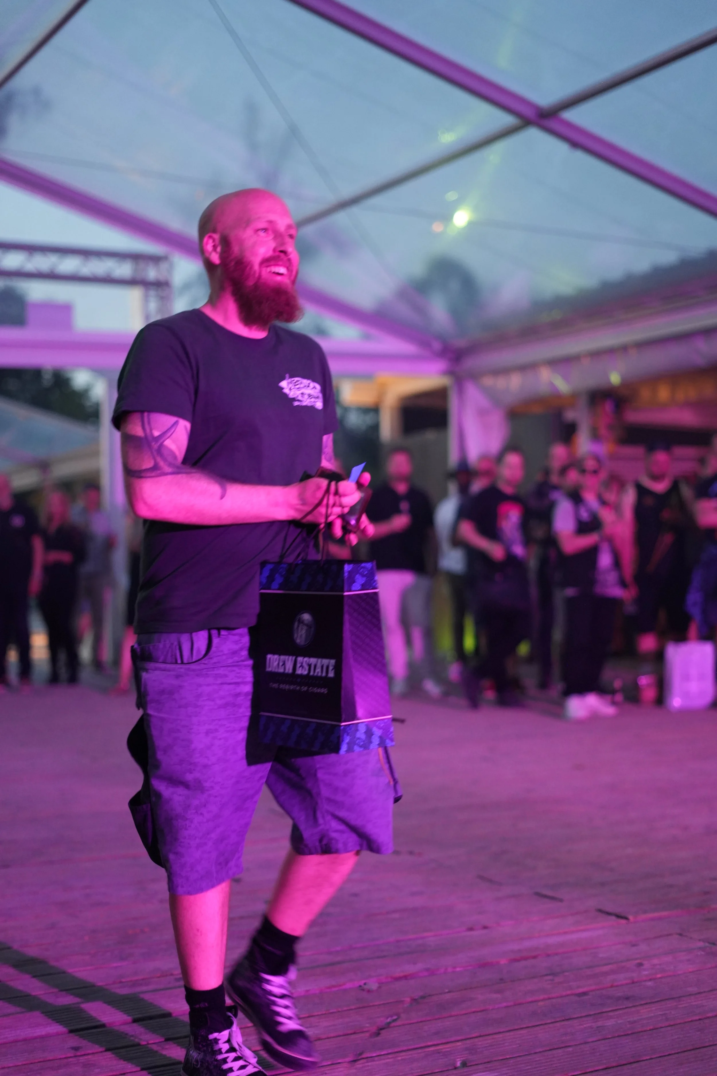 A man with a beard and tattoos, wearing a black T-shirt and shorts, standing under a glass canopy with pink and purple lights at an outdoor event, holding a black bag with 