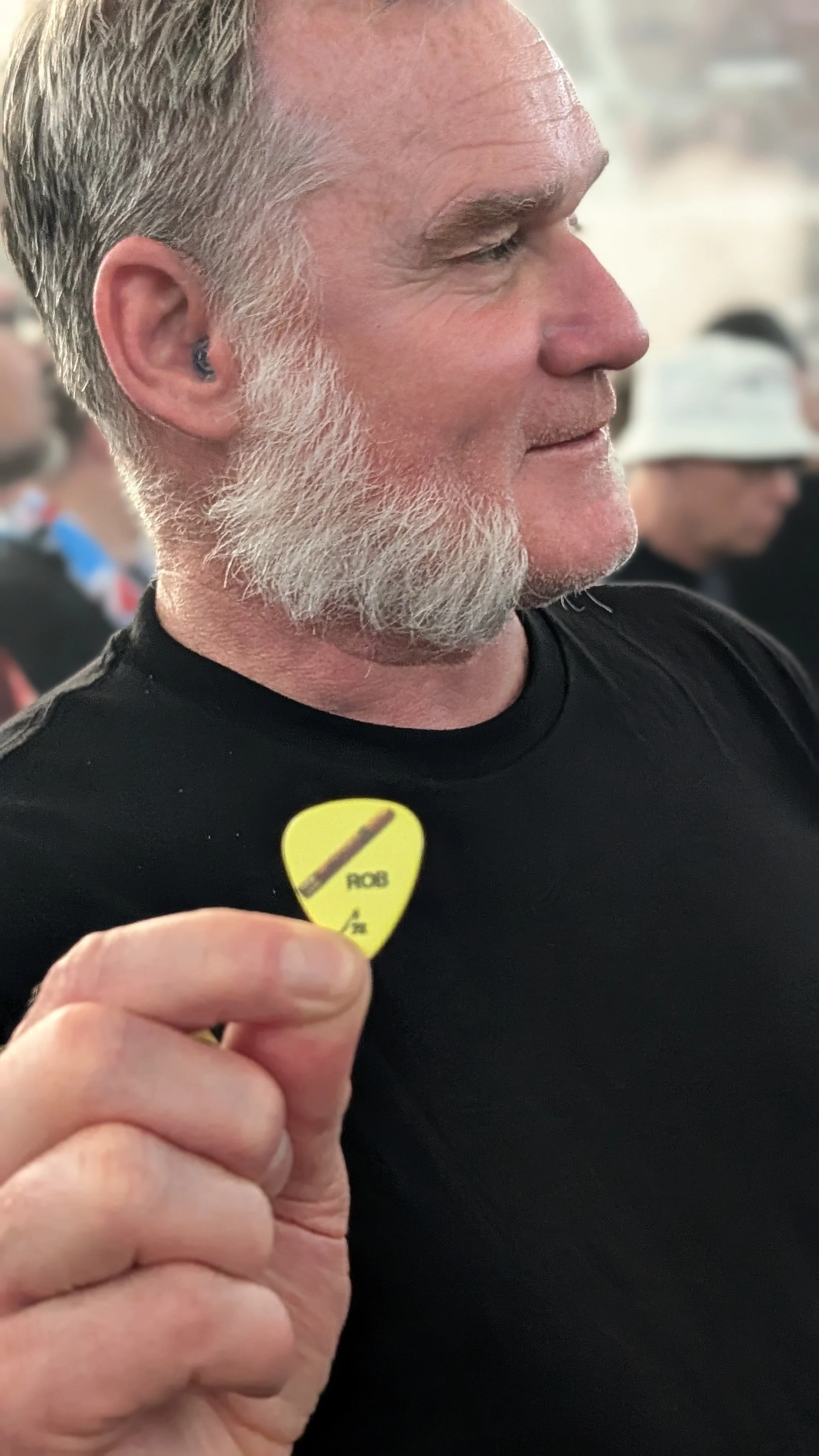 A man with gray hair and a beard holding a yellow guitar pick with a black pencil logo and the word 'PENCIL' on it, near his chest.