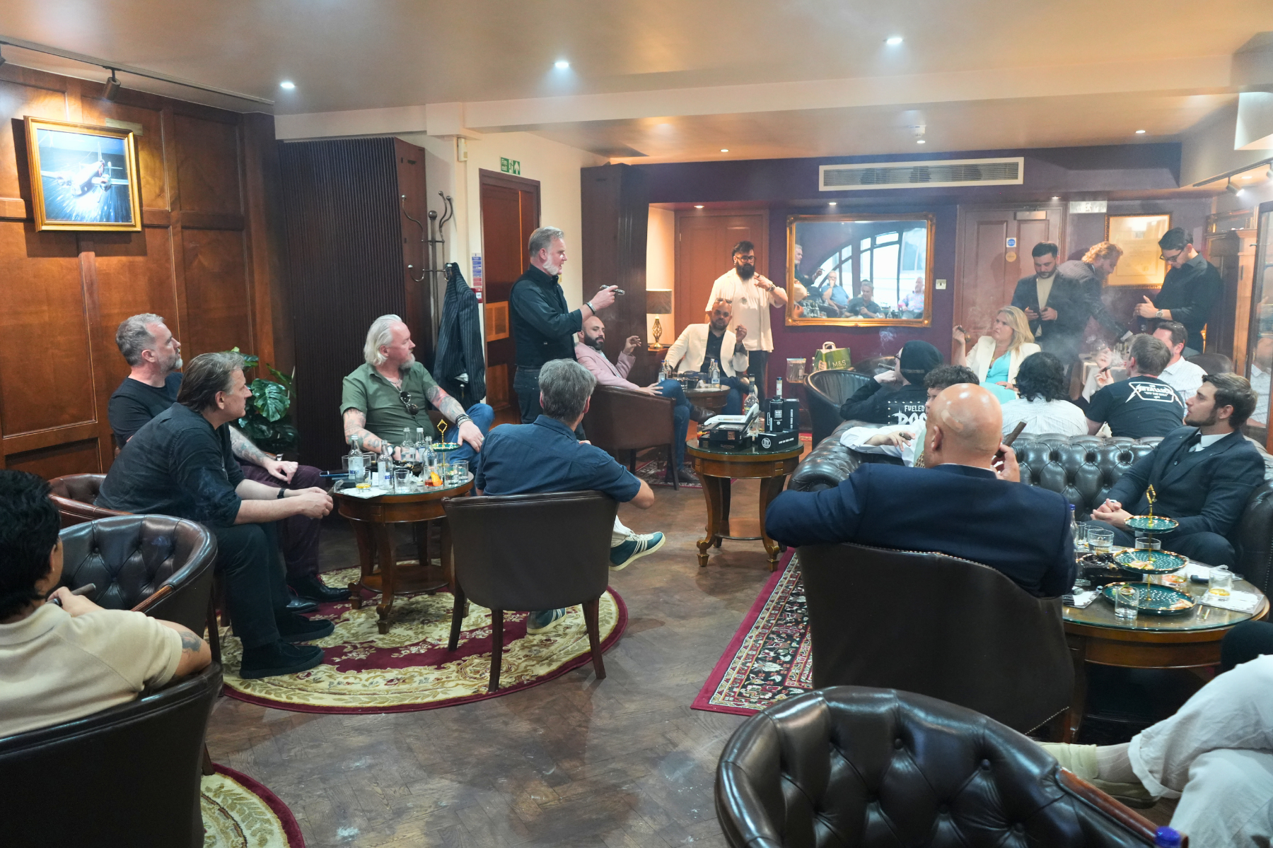 A group of people in a cozy, wood-paneled room with leather furniture, gathered for a social event or celebration. Some are toasting, sipping drinks, and chatting, with a few standing and others seated around tables with snacks and drinks. A man stan