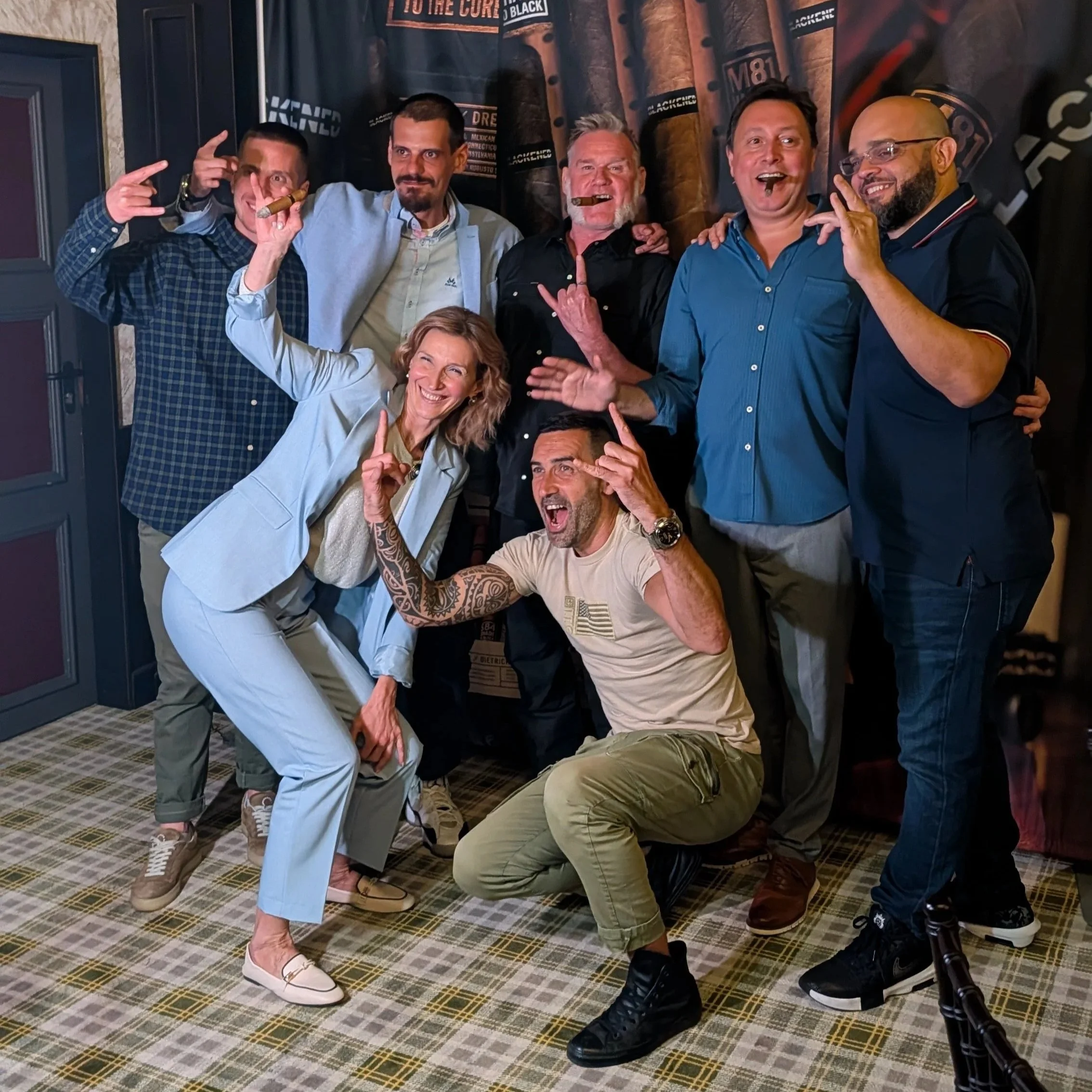 A group of seven smiling adults posing playfully in a restaurant, with some pointing or making gestures, behind a wall with cigar-themed decor.