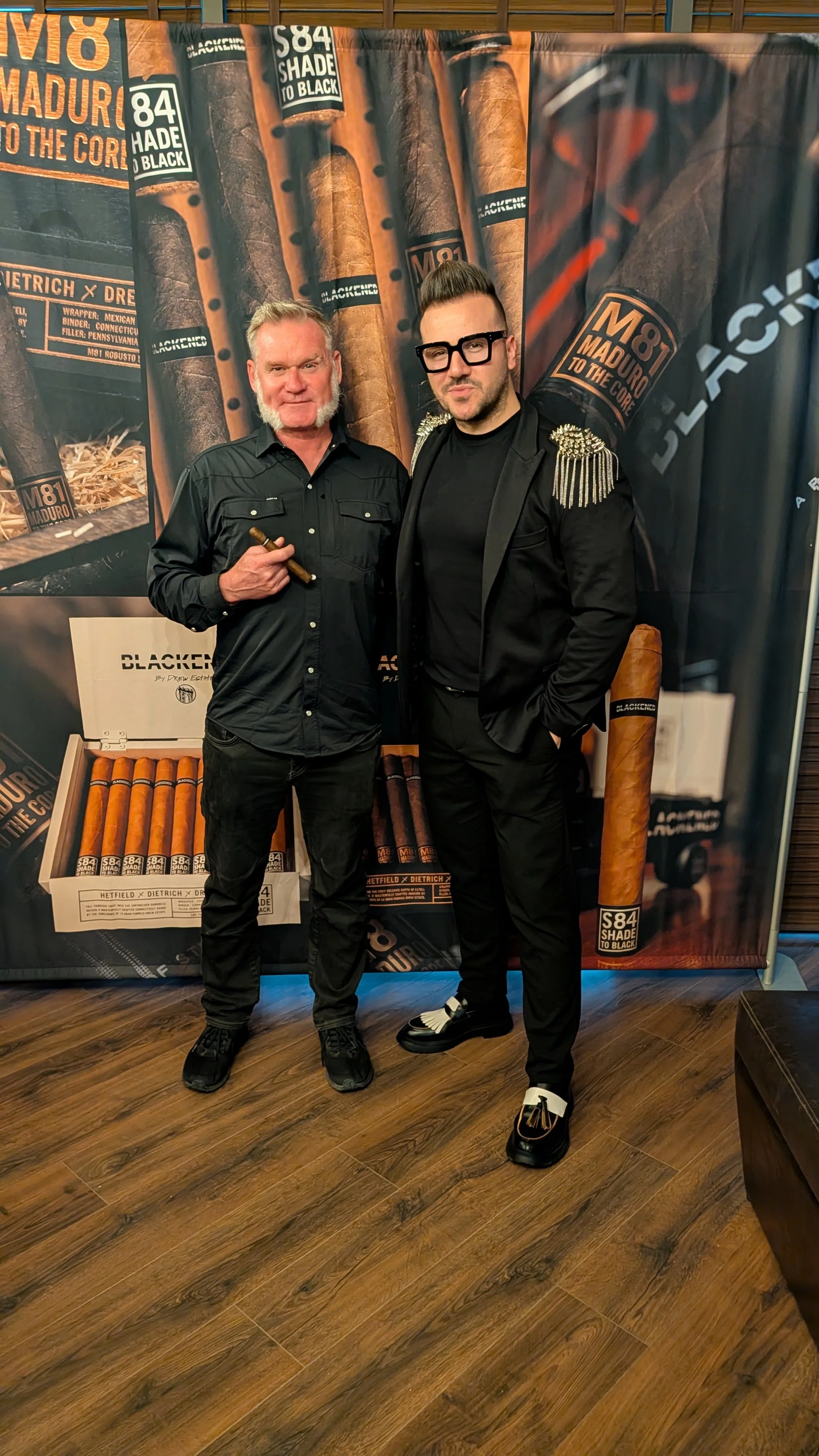 Two men standing in front of a background with cigar images and branding, one holding a cigar. The man on the left has gray hair and a beard, wearing a black button-up shirt and black pants. The man on the right has dark hair styled upward, wearing g