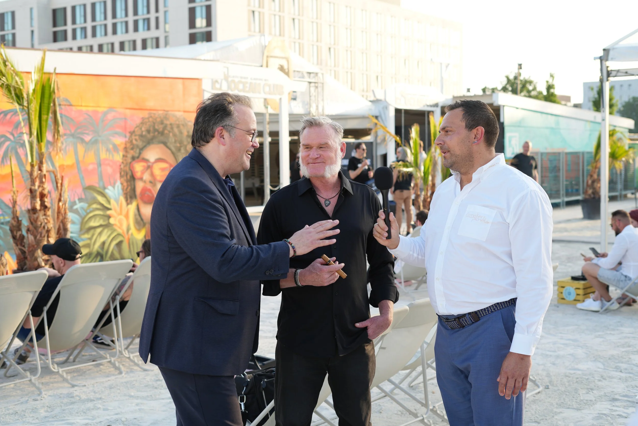 Three men are having a conversation outdoors at a daytime event, with lounge chairs and a colorful mural in the background.