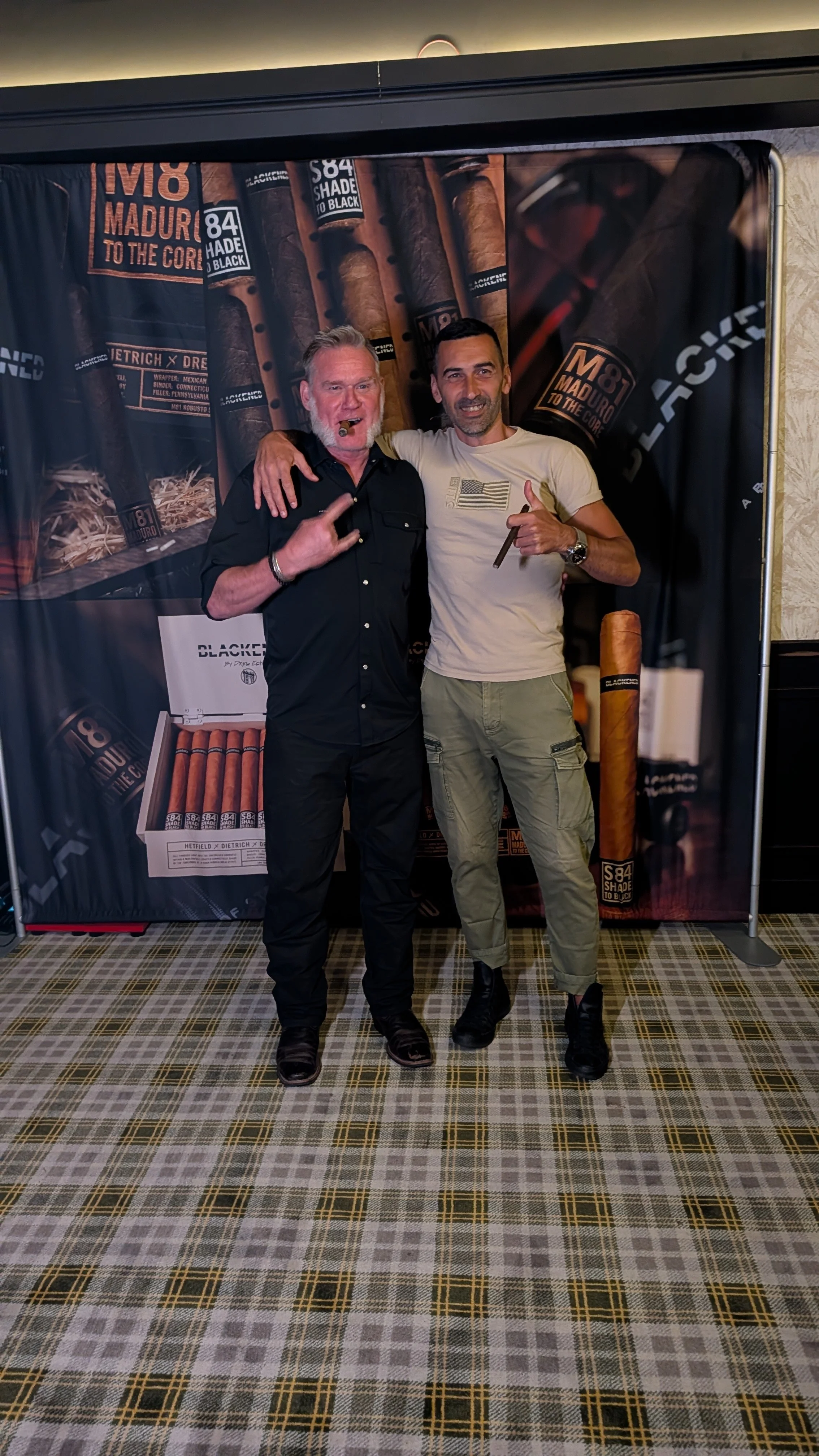 Two men standing together in front of a cigar-themed backdrop, one smoking a cigar and making a peace sign with his hand, while the other is pointing at his own cigar and giving a thumbs up.