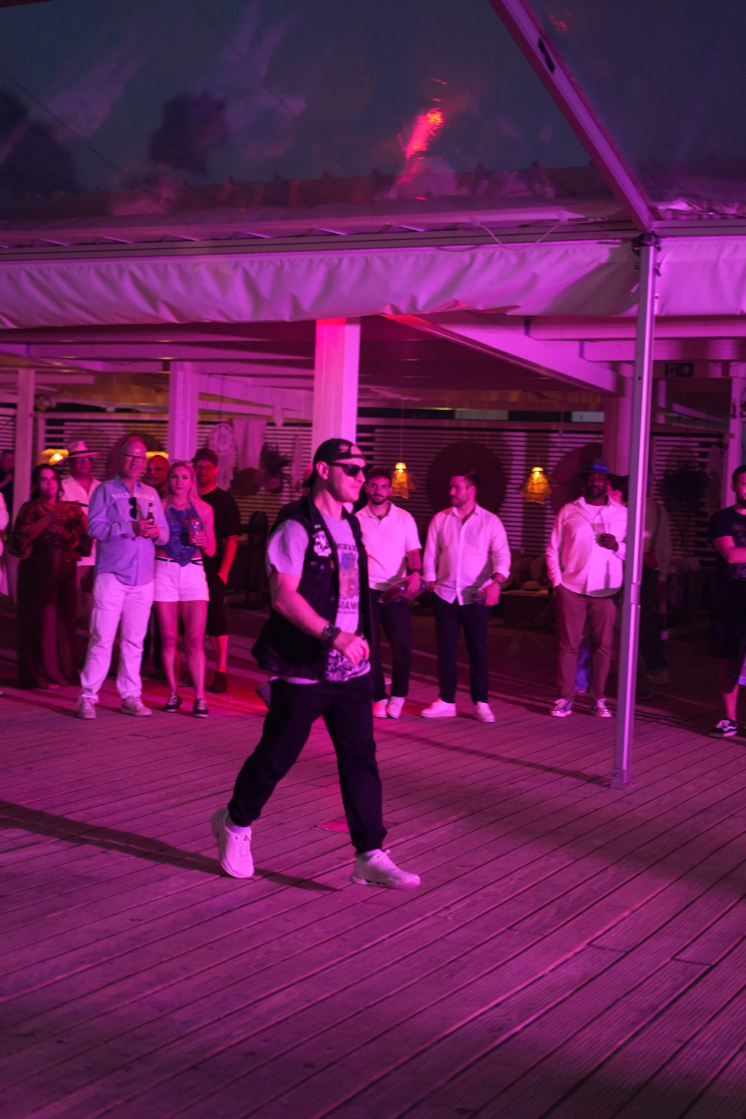 People gathered under a pavilion with pink lighting, some holding drinks, with a man in the forefront walking. The structure is made of wooden flooring, columns, and a tent-like cover overhead.