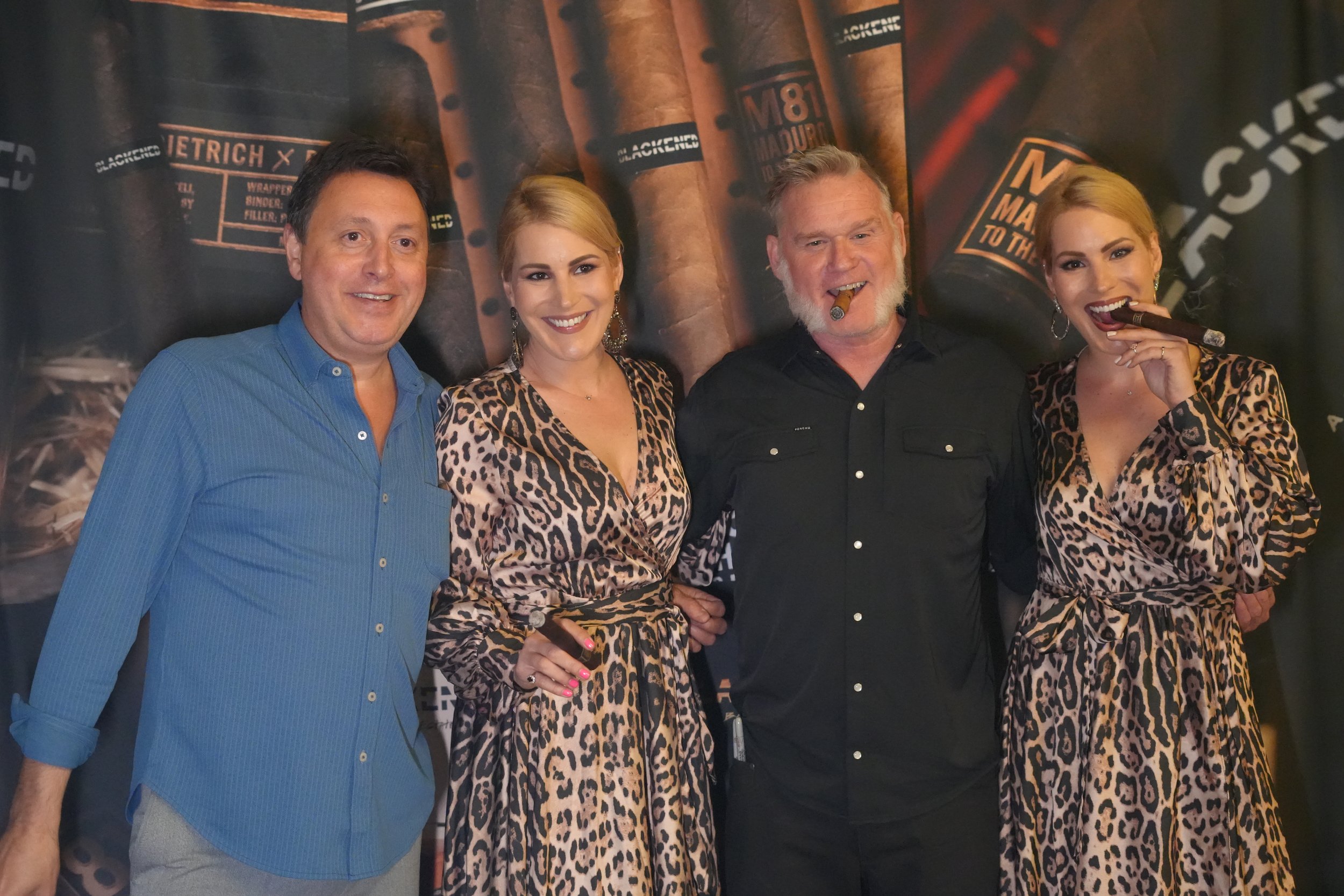Four people posing together indoors, two women and two men, with a background featuring cigars and cigar-related branding. The women are wearing leopard print outfits, and the men are wearing dark and blue shirts. All are smiling, and one woman is ho