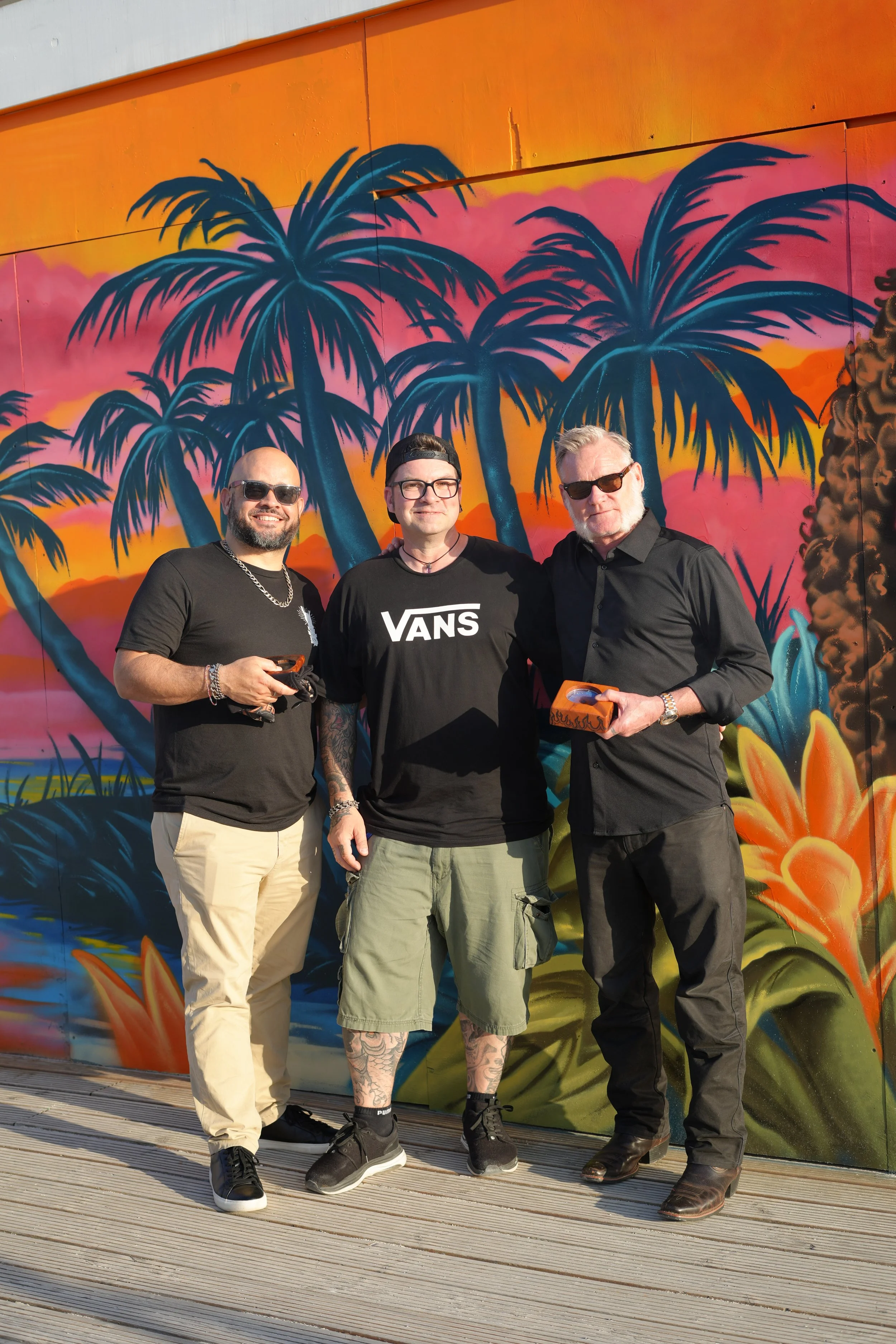 Three men stand in front of a colorful mural featuring palm trees and a sunset. The man on the left is wearing sunglasses, a black t-shirt, beige pants, and has a silver chain necklace. The man in the middle is wearing glasses, a black Vans t-shirt, 