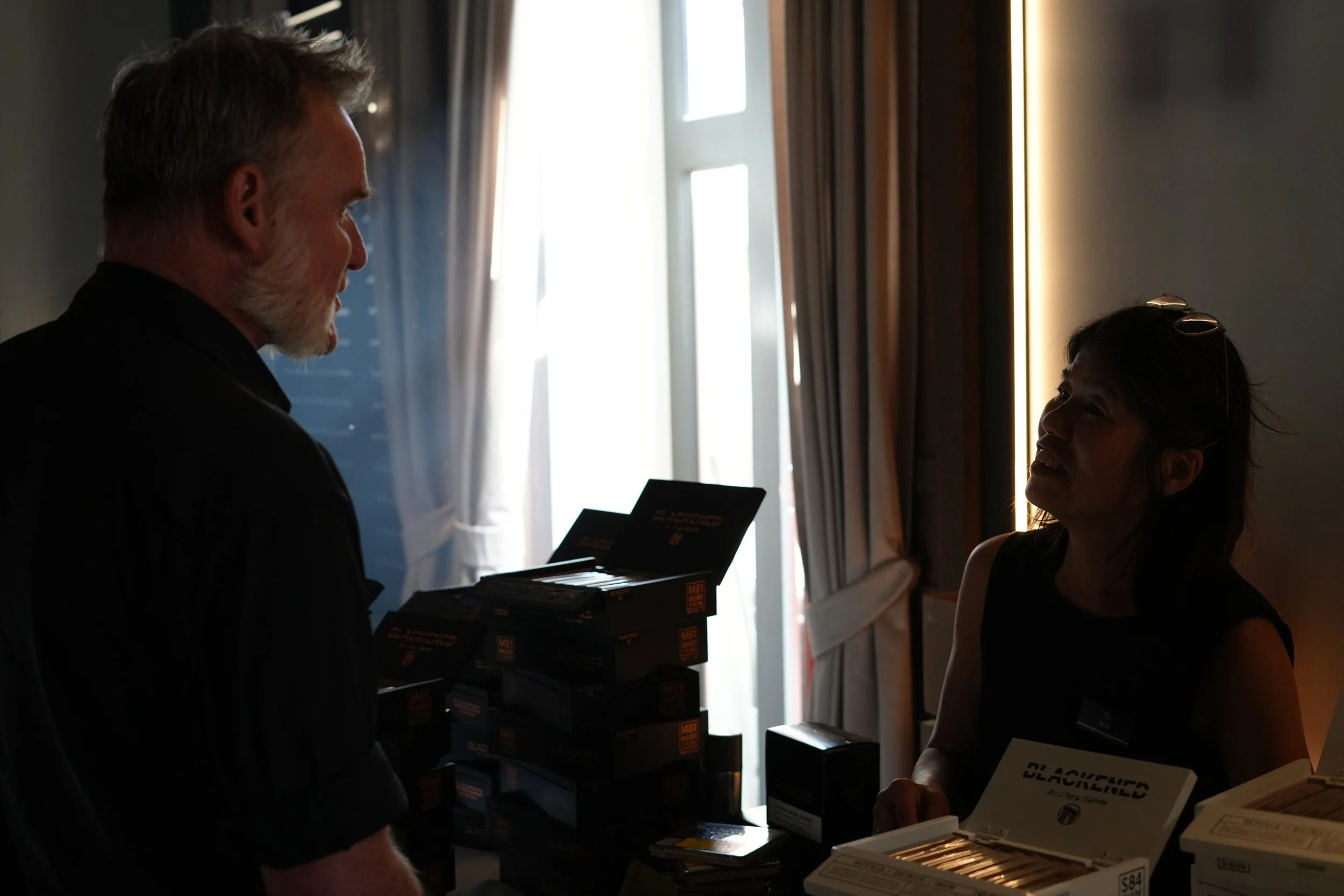 A man with gray hair and beard talking to a woman with sunglasses on her head. They are standing near a table with boxes and pamphlets, with light coming from a window behind them.