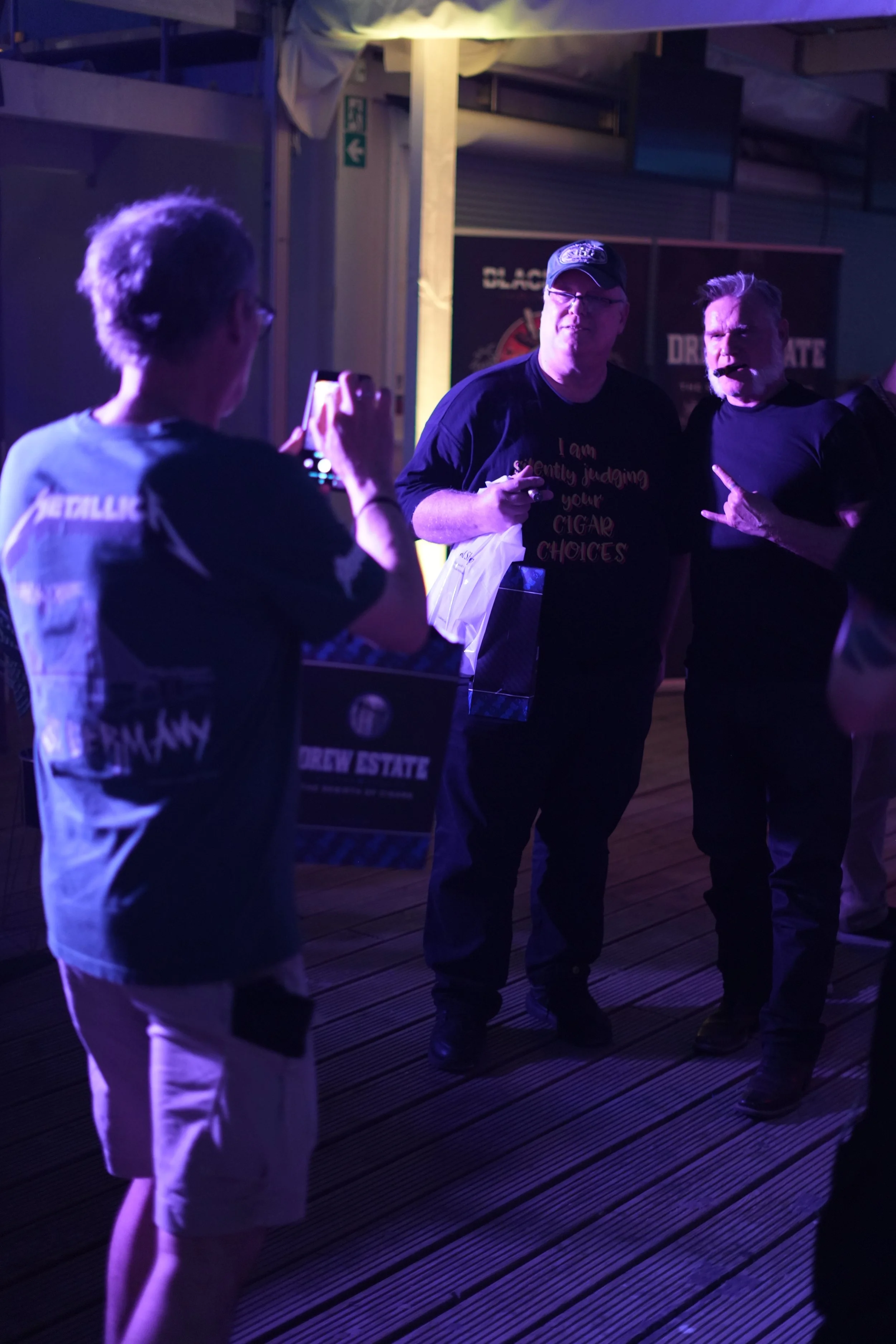 Three men and one woman standing at a gathering or party, with one woman taking a photo of two of the men. The setting appears to be indoors or in a covered area, with some banners or signs in the background. The lighting is dim with purple and yello