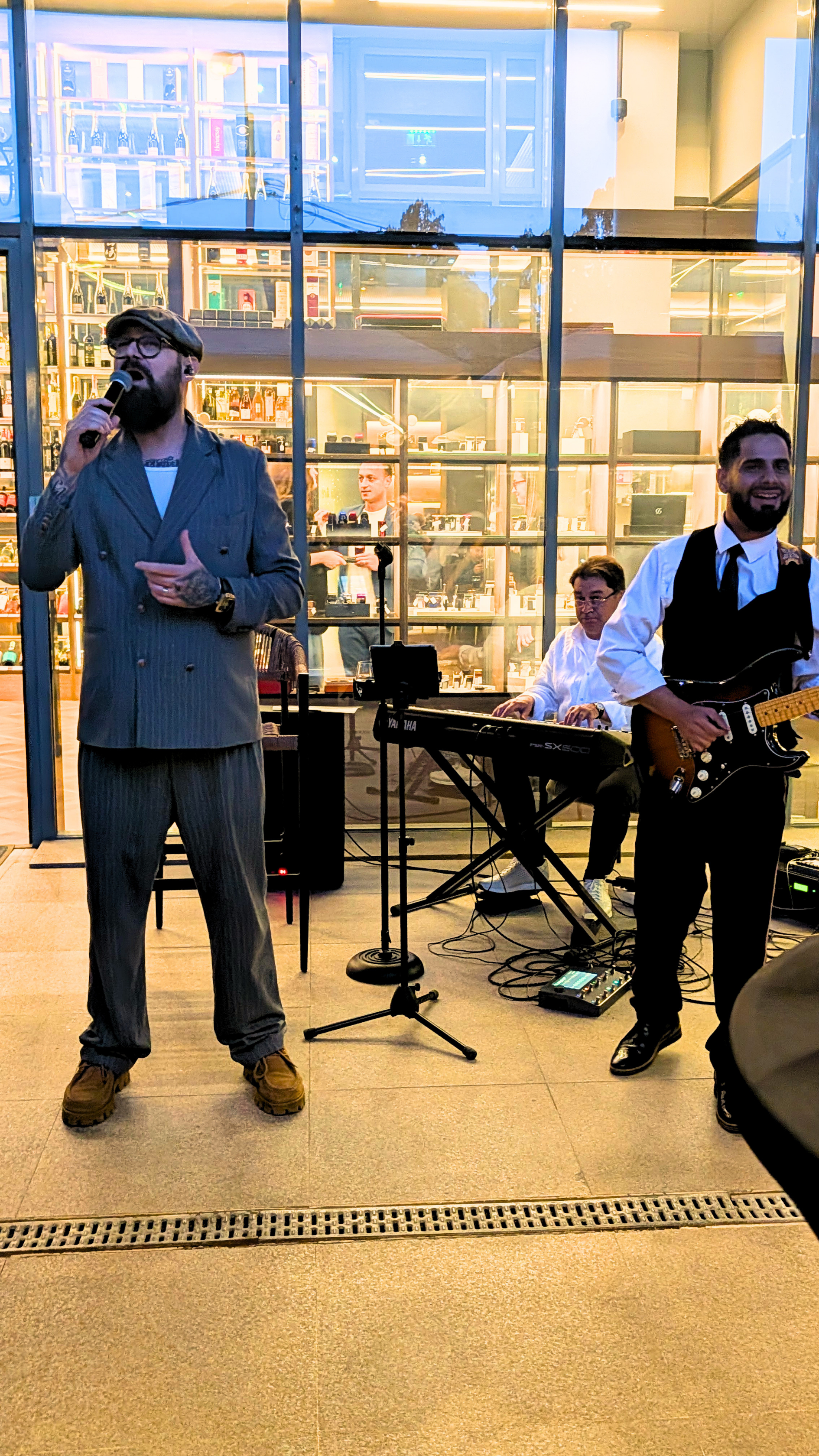 A band performs in front of a large glass window at an indoor venue. The band includes a singer with a beard and glasses, a keyboard player, and a guitarist. The window reveals shelves with bottles and books inside the building.