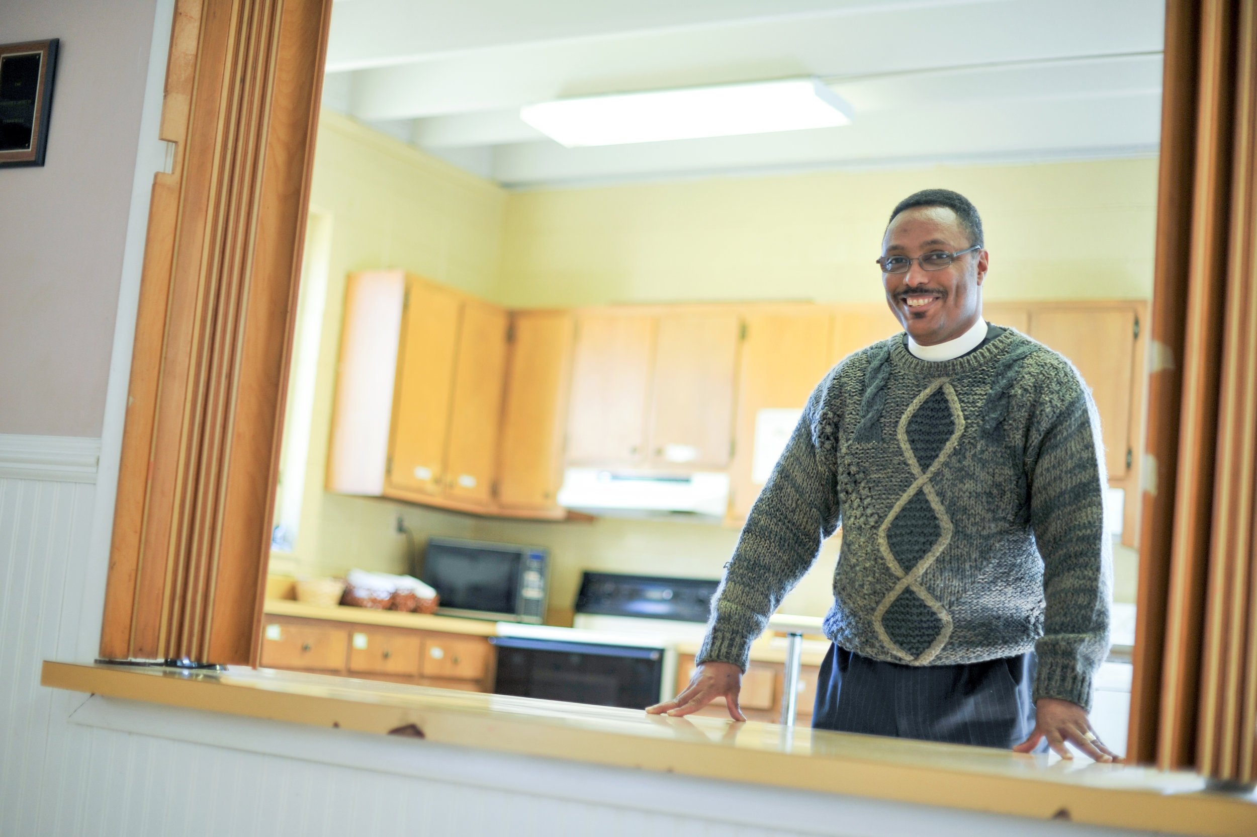 Man in a sweater smiling in a kitchen setting with wooden cabinets and countertop.