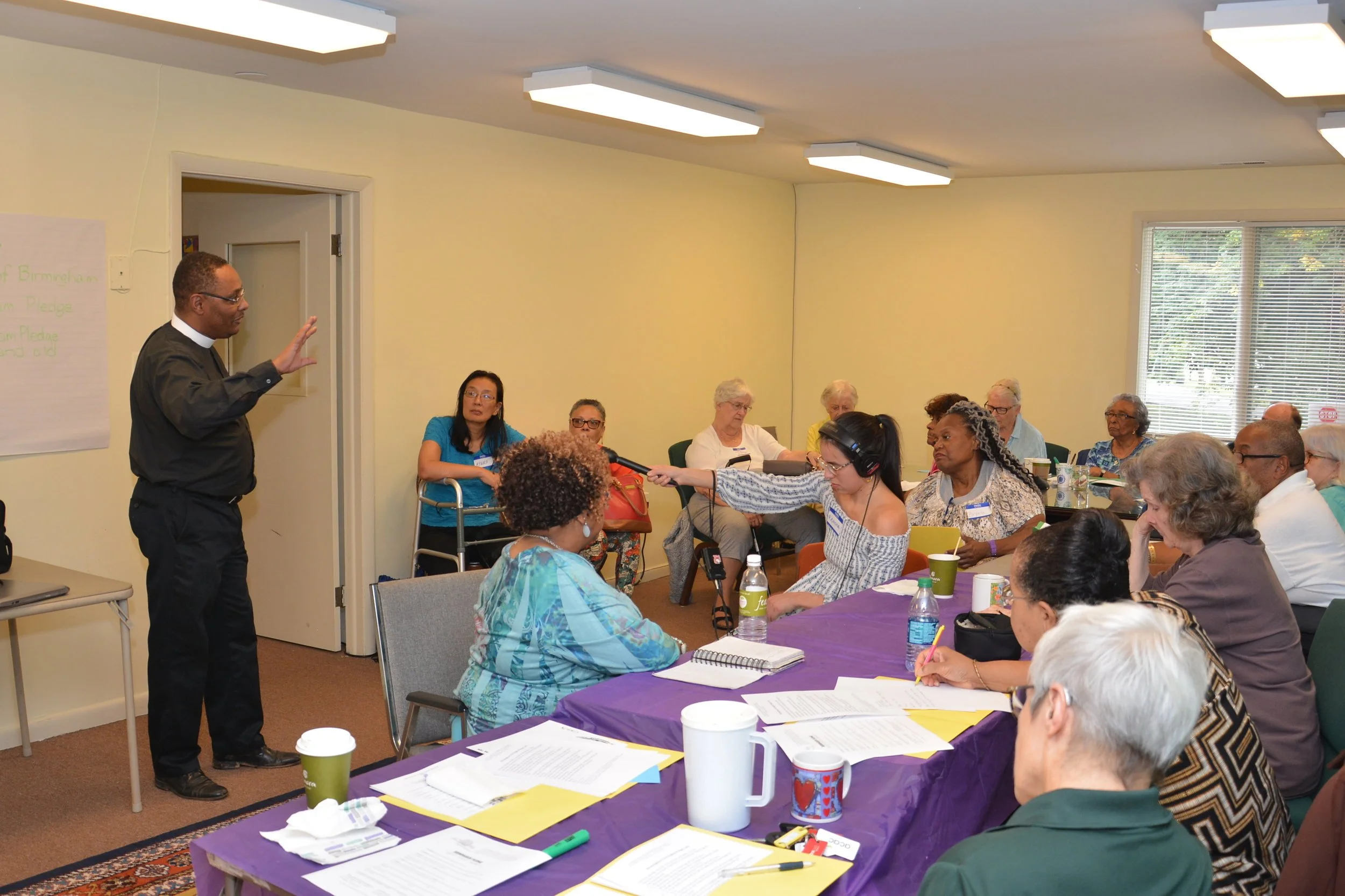 A group of elderly individuals attending a seminar in a classroom setting. A man is speaking at the front of the room, while the attendees are seated around a table covered with papers, cups, and handouts. A woman with headphones is holding a microphone, possibly conducting an interview or recording.