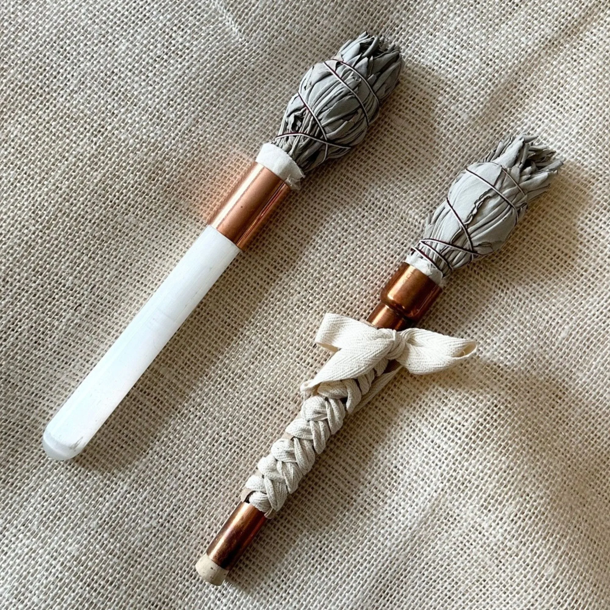 Close-up of two bundle of sage smudge sticks tied with black rubber bands, lying on a textured beige surface.