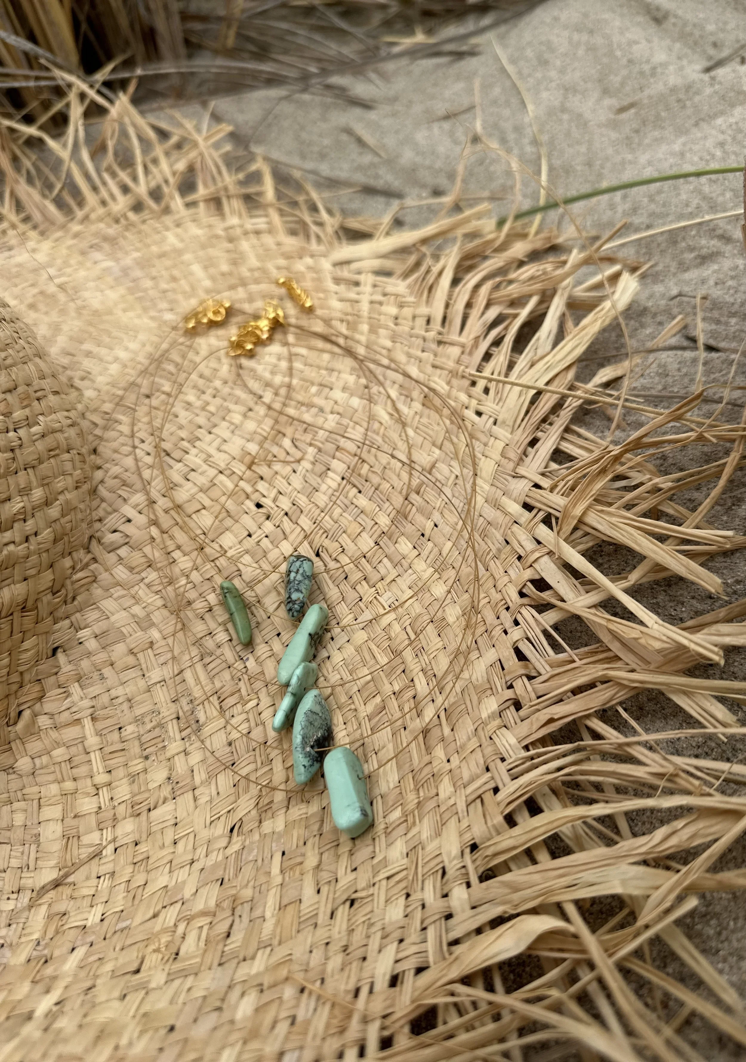 A woven straw hat on a stone surface with small turquoise stones and gold decorative beads placed on it, surrounded by dry grass or straw.