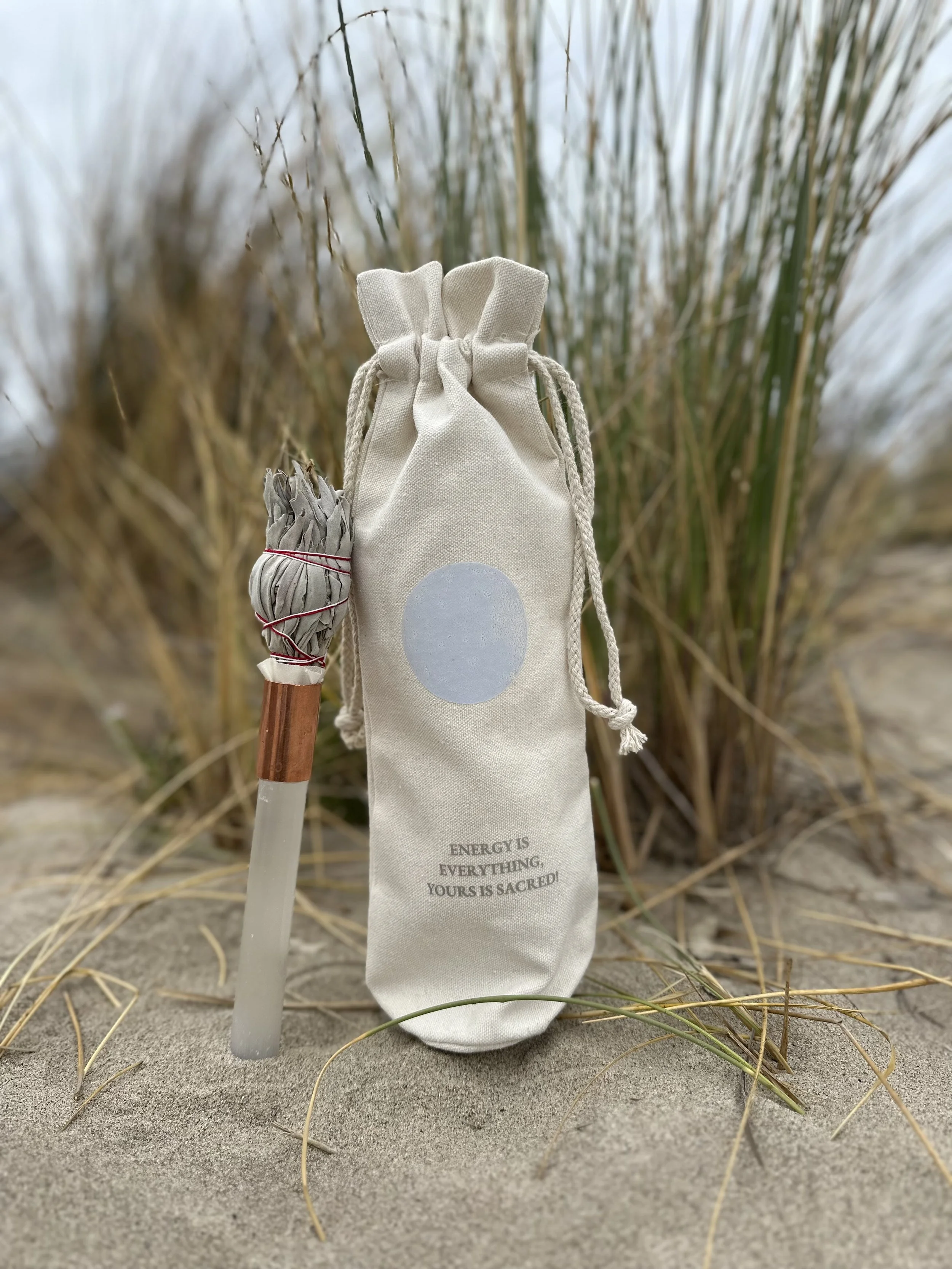 A small white drawstring bag with a gray circle and the quote "Energy is everything. Yours is sacred!" printed on it, placed on the sand among tall beach grasses.