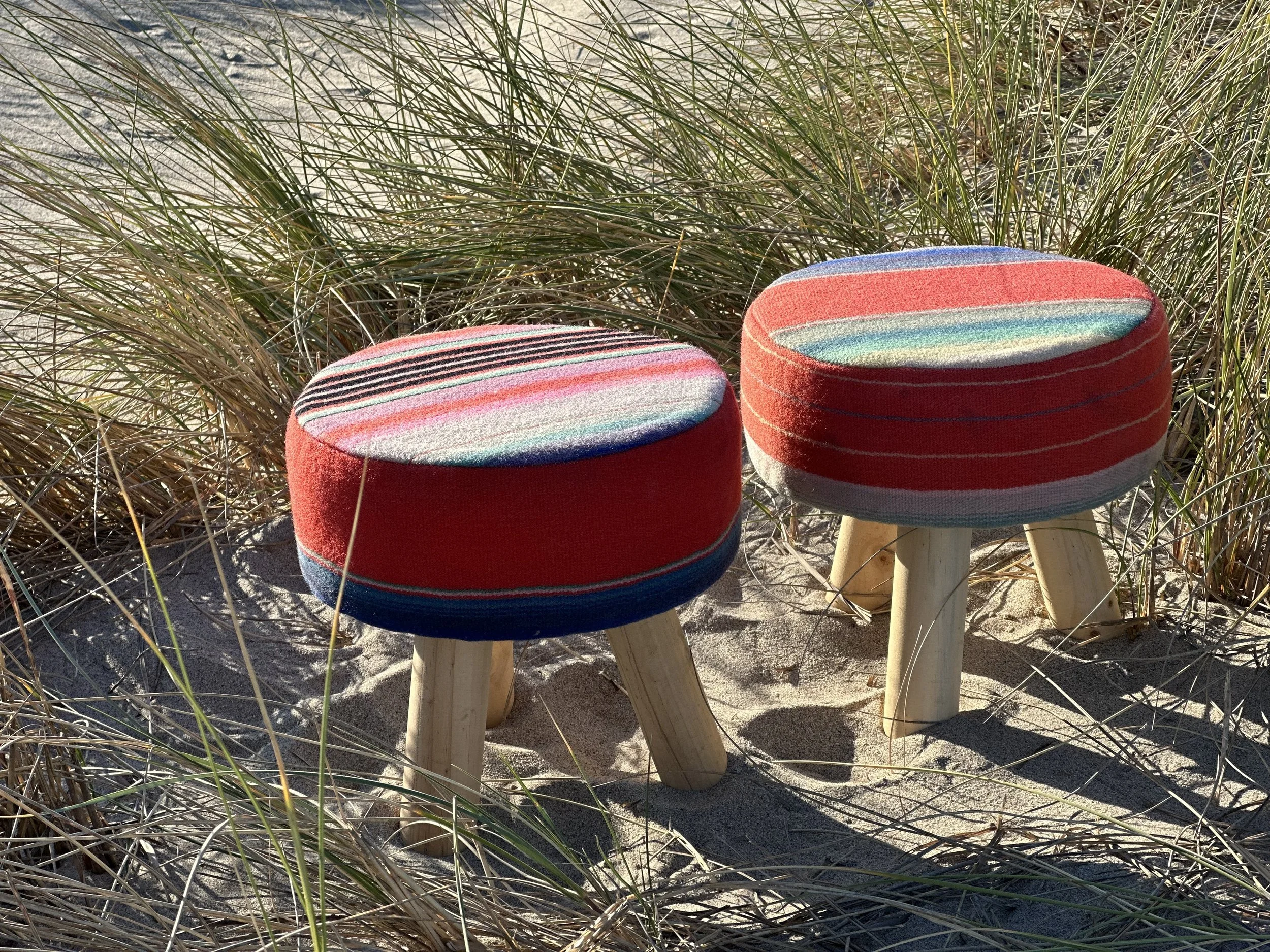 Two round cushioned seats with colorful striped fabric, placed outdoors among sandy terrain and grass dunes.