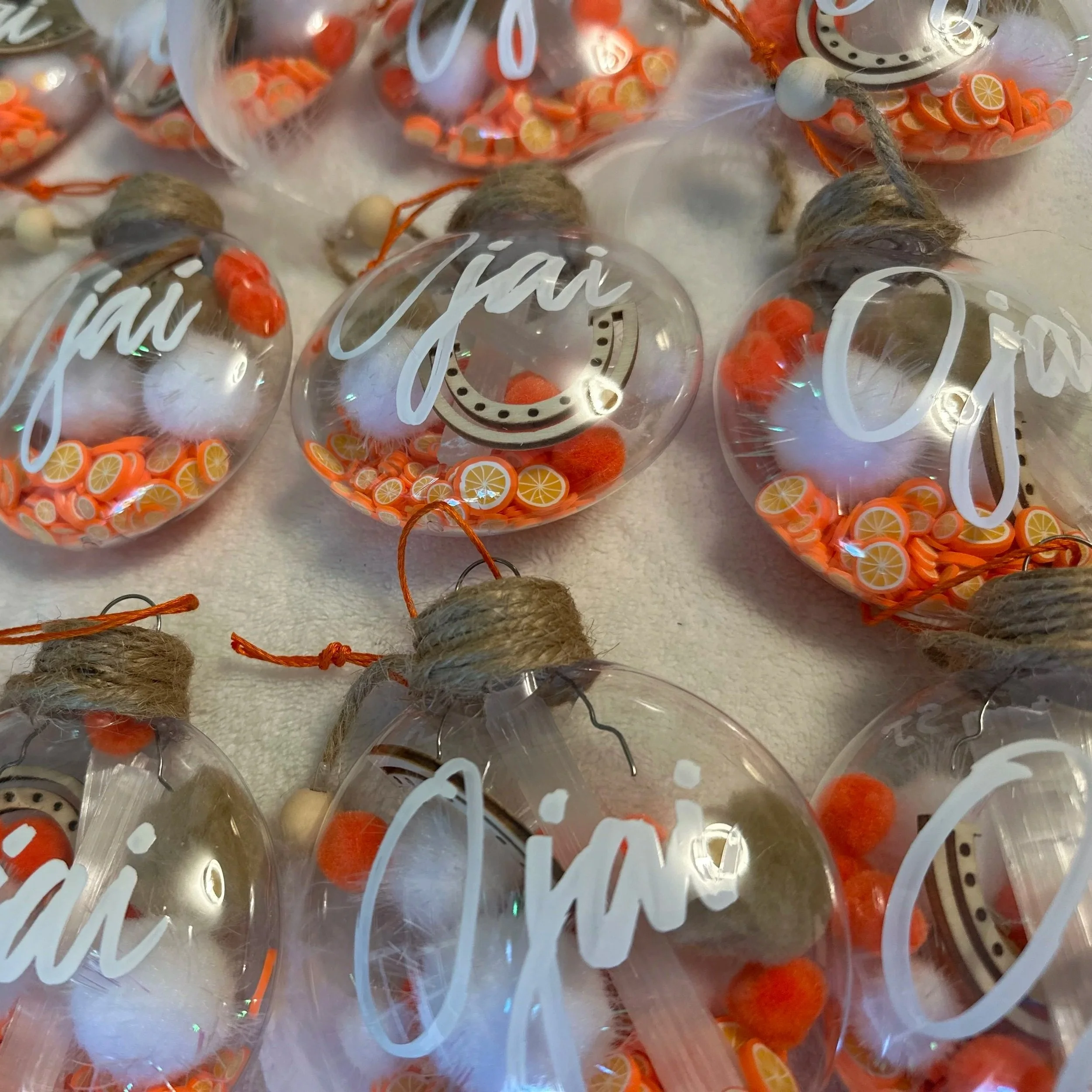 Multiple clear plastic ornaments filled with small orange and yellow lemon slice decorations, with some ornaments containing fluffy white material, and with white and orange string hanging from some.