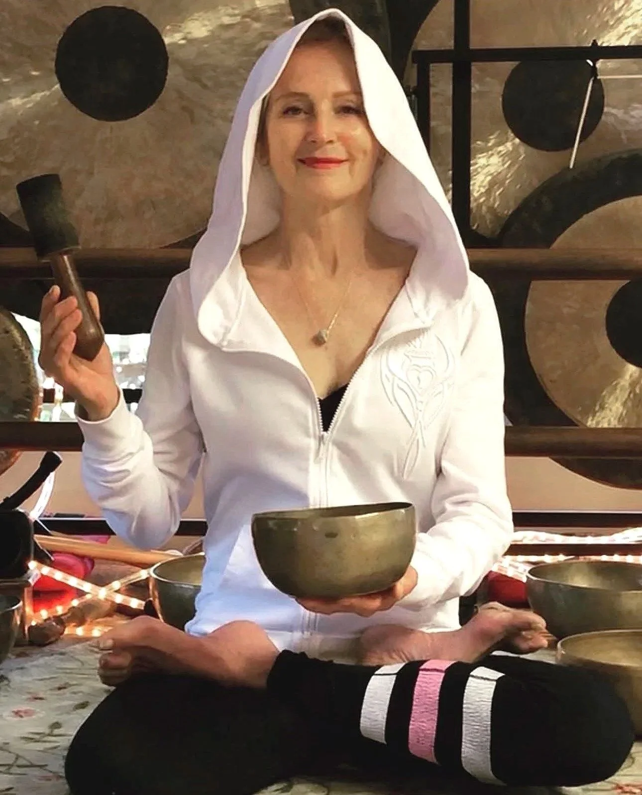 A woman in a white hoodie sitting cross-legged at a table with singing bowls, holding a bowl in her left hand and a mallet in her right hand, smiling in a room with a musical instrument wall decoration in the background.
