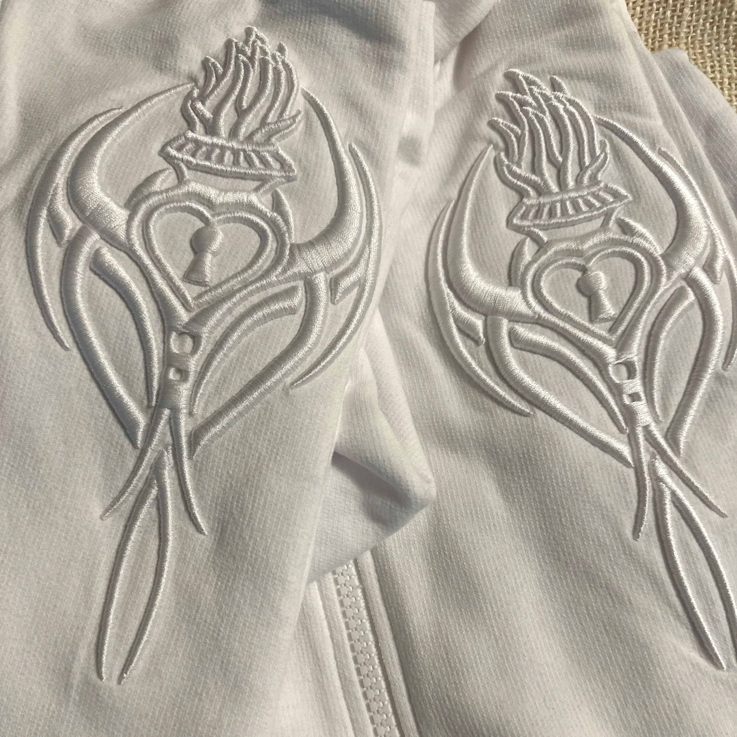 Close-up of a white fabric with intricate silver embroidery of floral and leaf patterns, featuring a white zipper at the bottom.