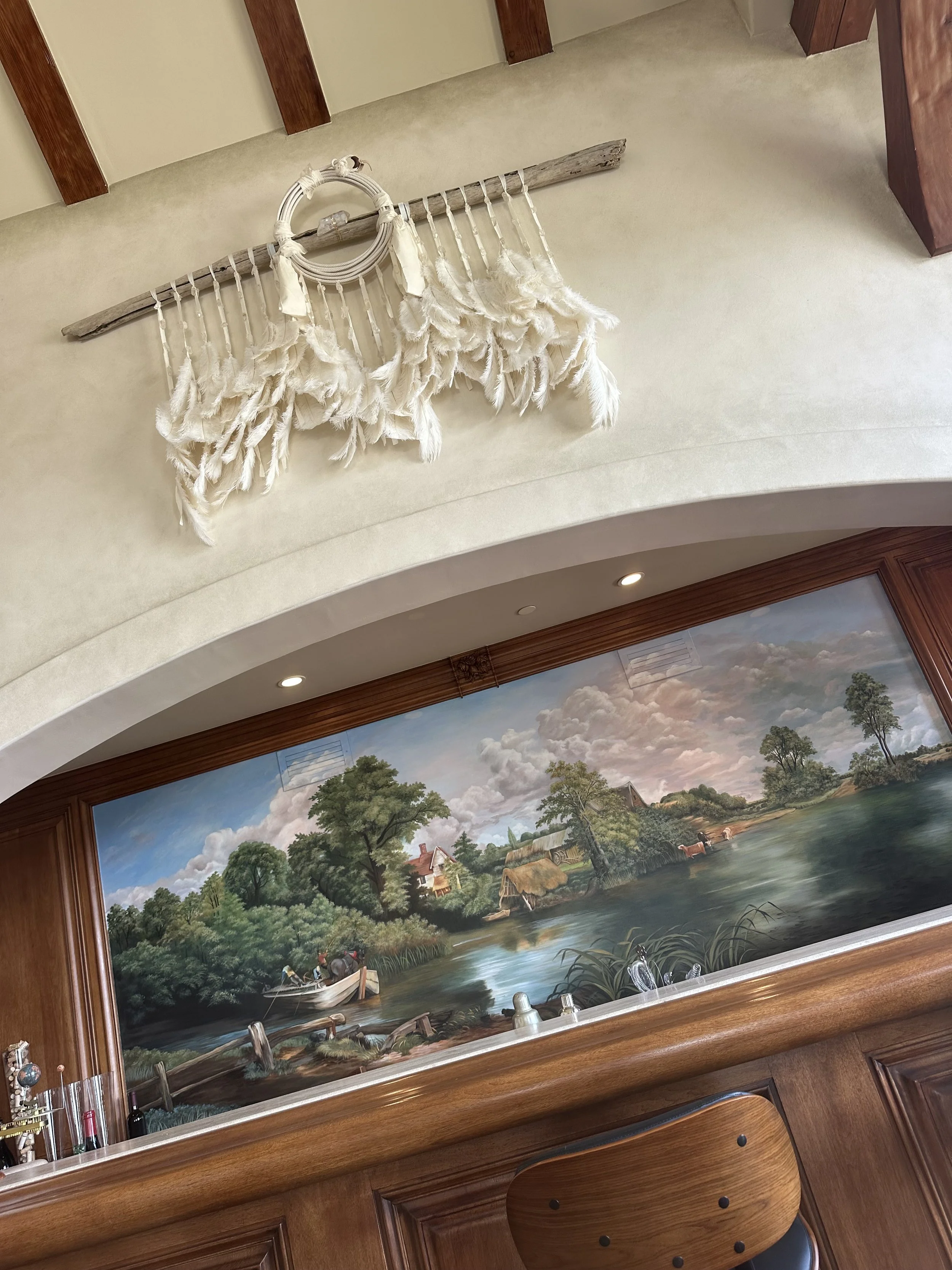 A mural painting of a rural landscape with trees, a river, and houses, situated behind a wooden bar with a decorative mirror and bar accessories.