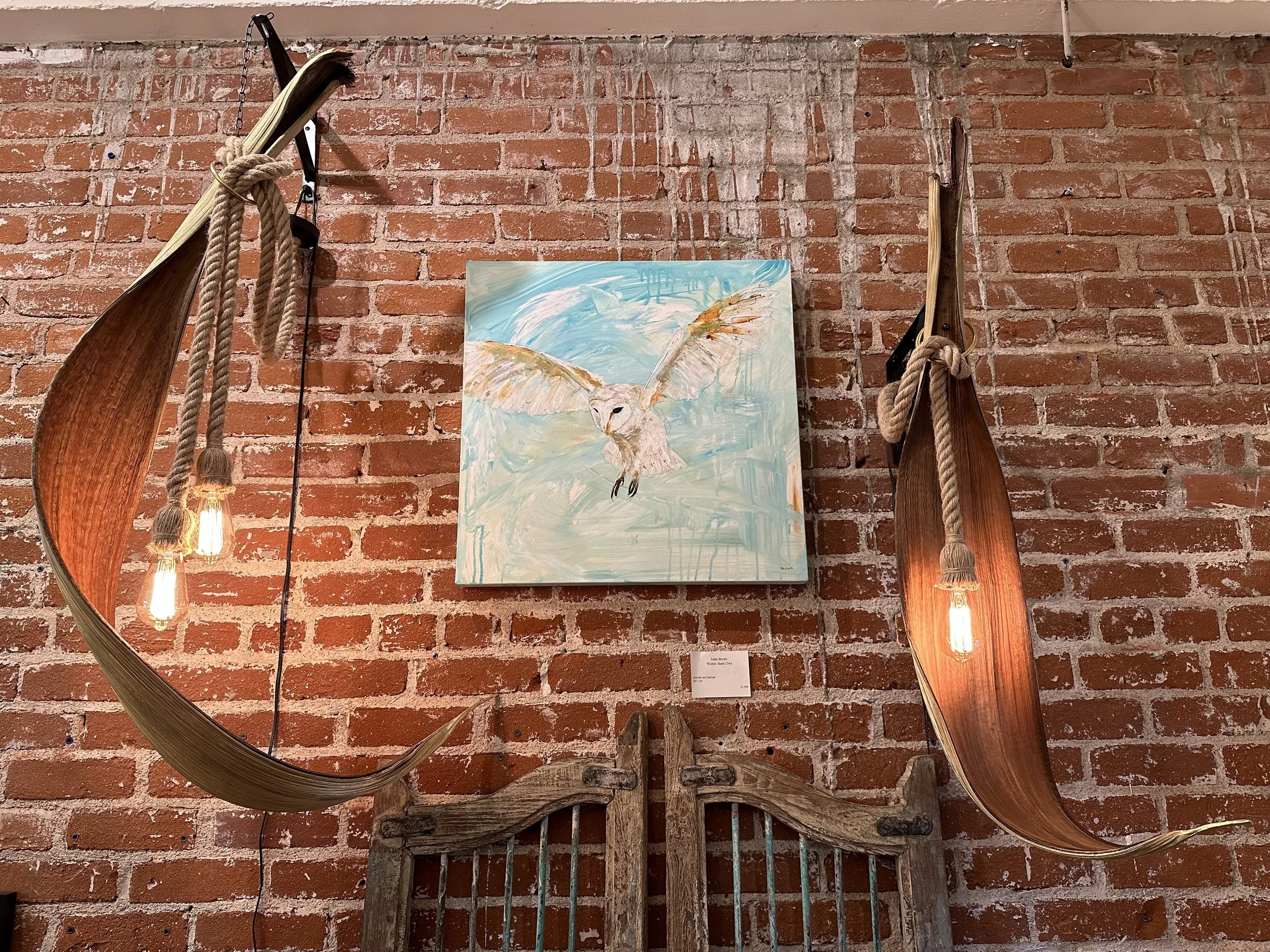 Art display featuring two long wooden canoes with ropes, a colorful painting of an owl with open wings on a brick wall, and vintage wooden tools beneath. Hanging light bulbs provide lighting.