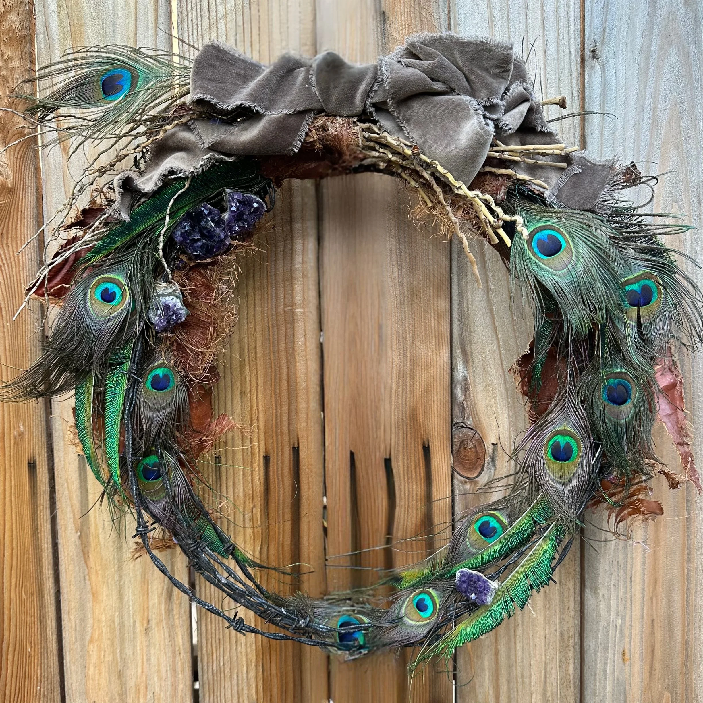 A decorative wreath made of peacock feathers, purple crystals, and a brown fabric, hanging on a wooden wall.