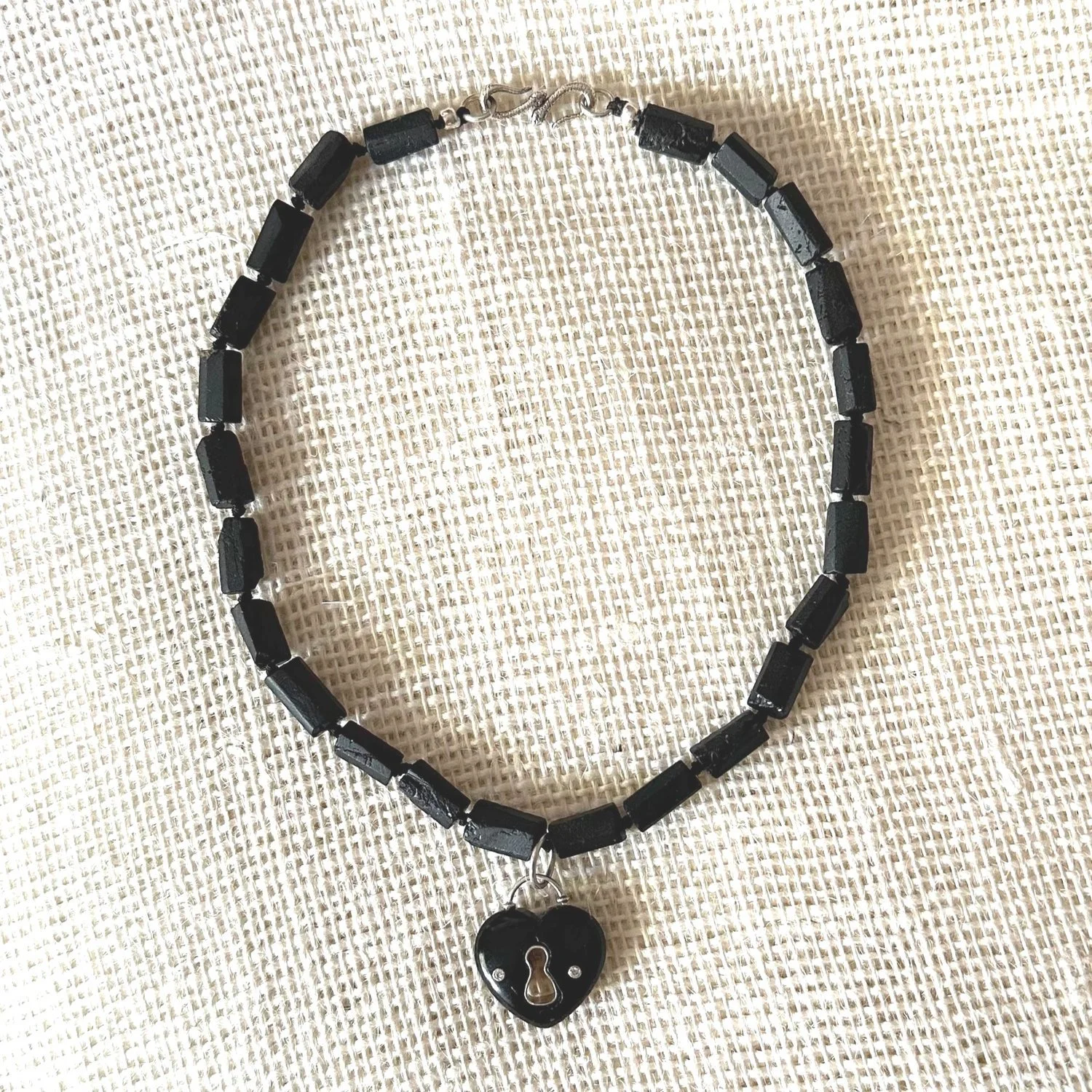 A black necklace with black rectangular beads and a black heart-shaped pendant featuring a keyhole design, laid on a beige textured fabric background.