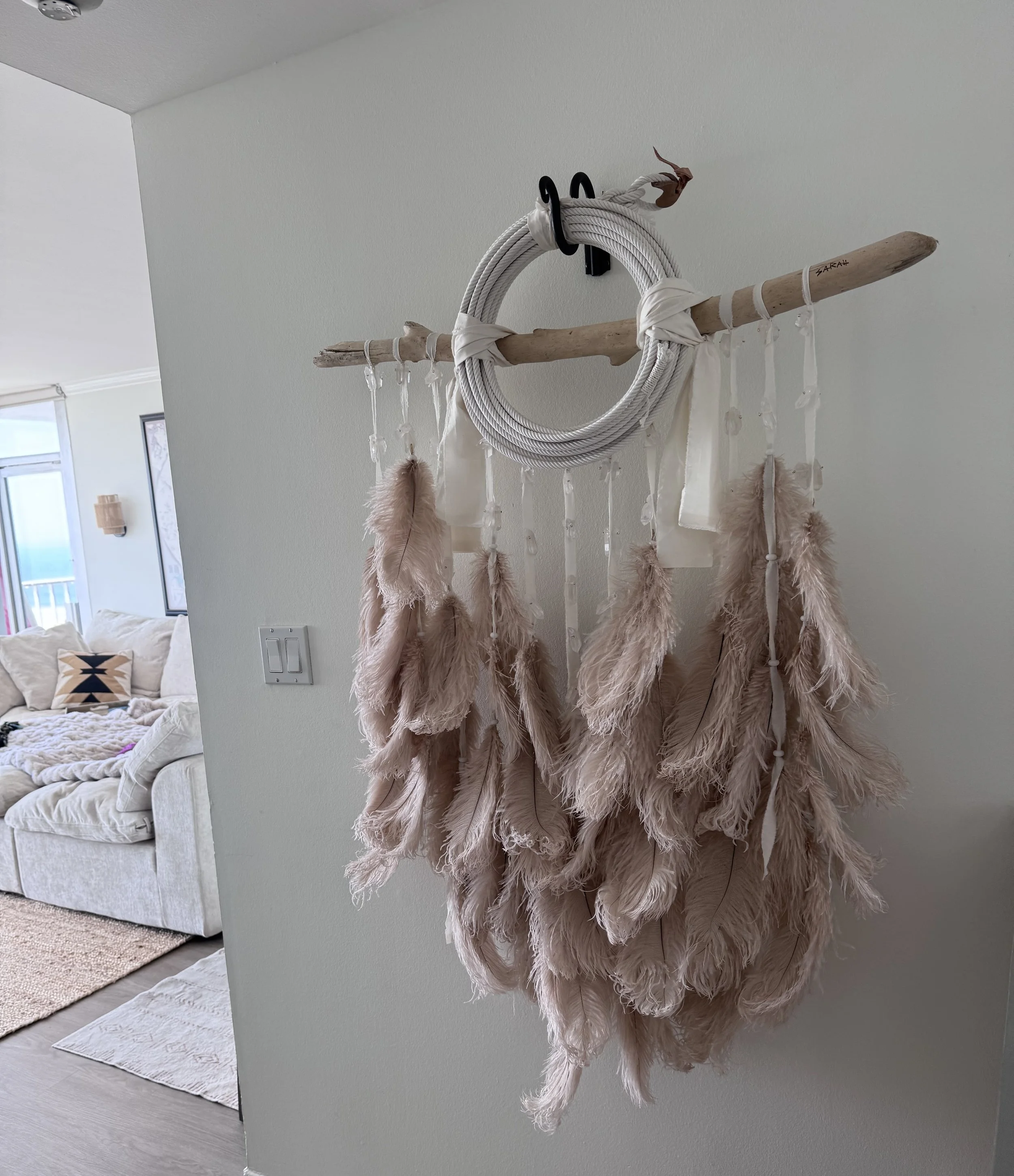 A cluster of pink feathered dreamcatchers hanging on a wooden stick, attached to a wall with black hooks, in a room with a white couch, a black and white pillow, and windows in the background.