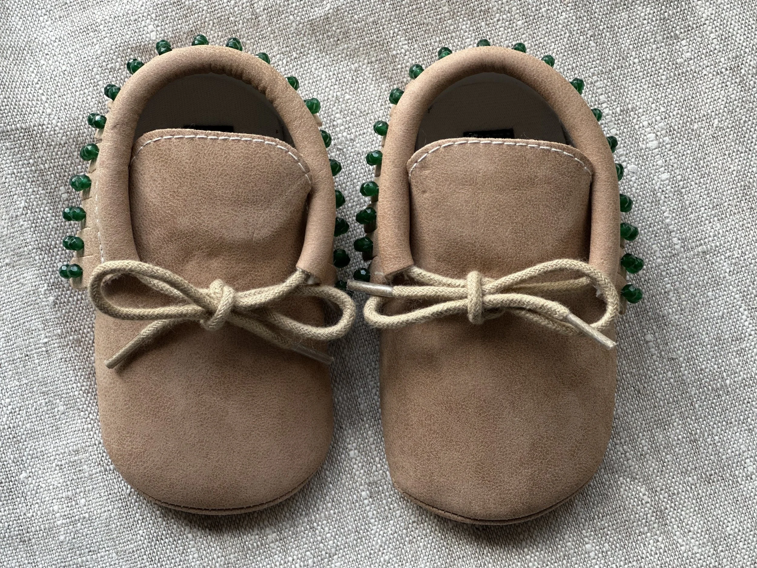 Pair of tan baby shoes with green bead embellishments around the edges, placed on a textured fabric surface.
