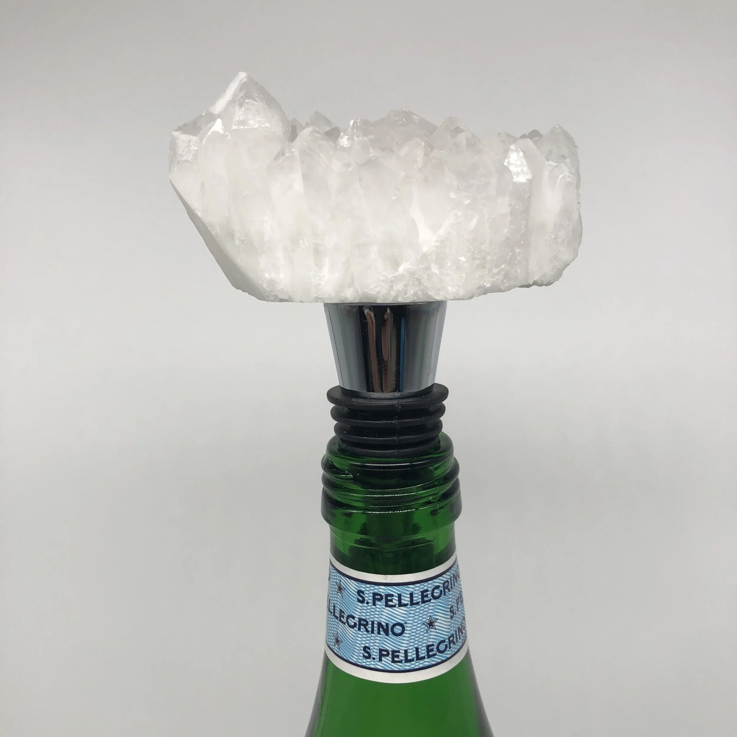 A close-up of a sparkling, clear, crystalline structure with a jagged appearance, mounted on a black and green spiral stem, with a blurred label reading "S. PELLEGRINO" in the background.