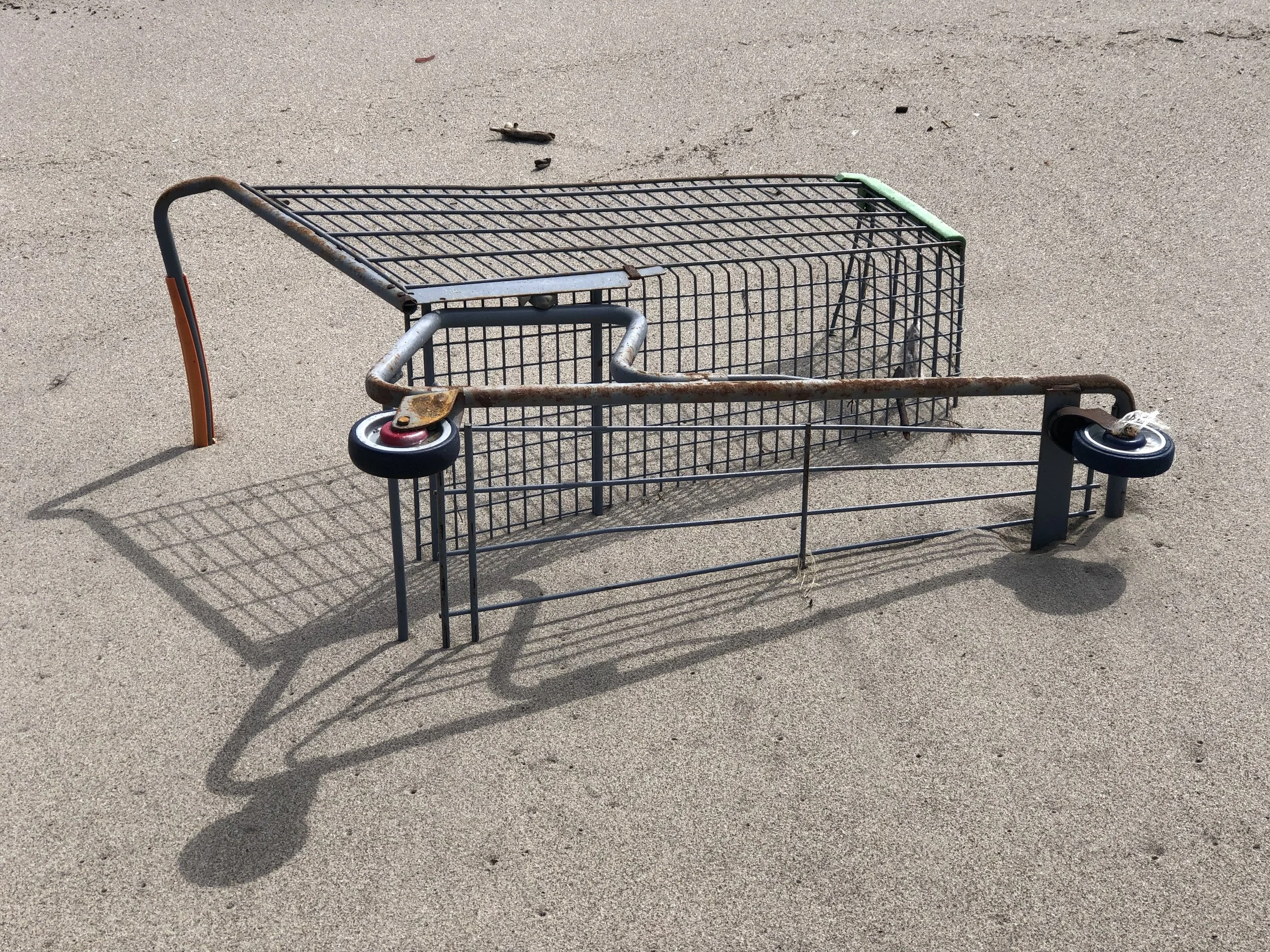 A rusty shopping cart lying on sandy ground with its castor wheel detached, and its shadow cast on the surface.
