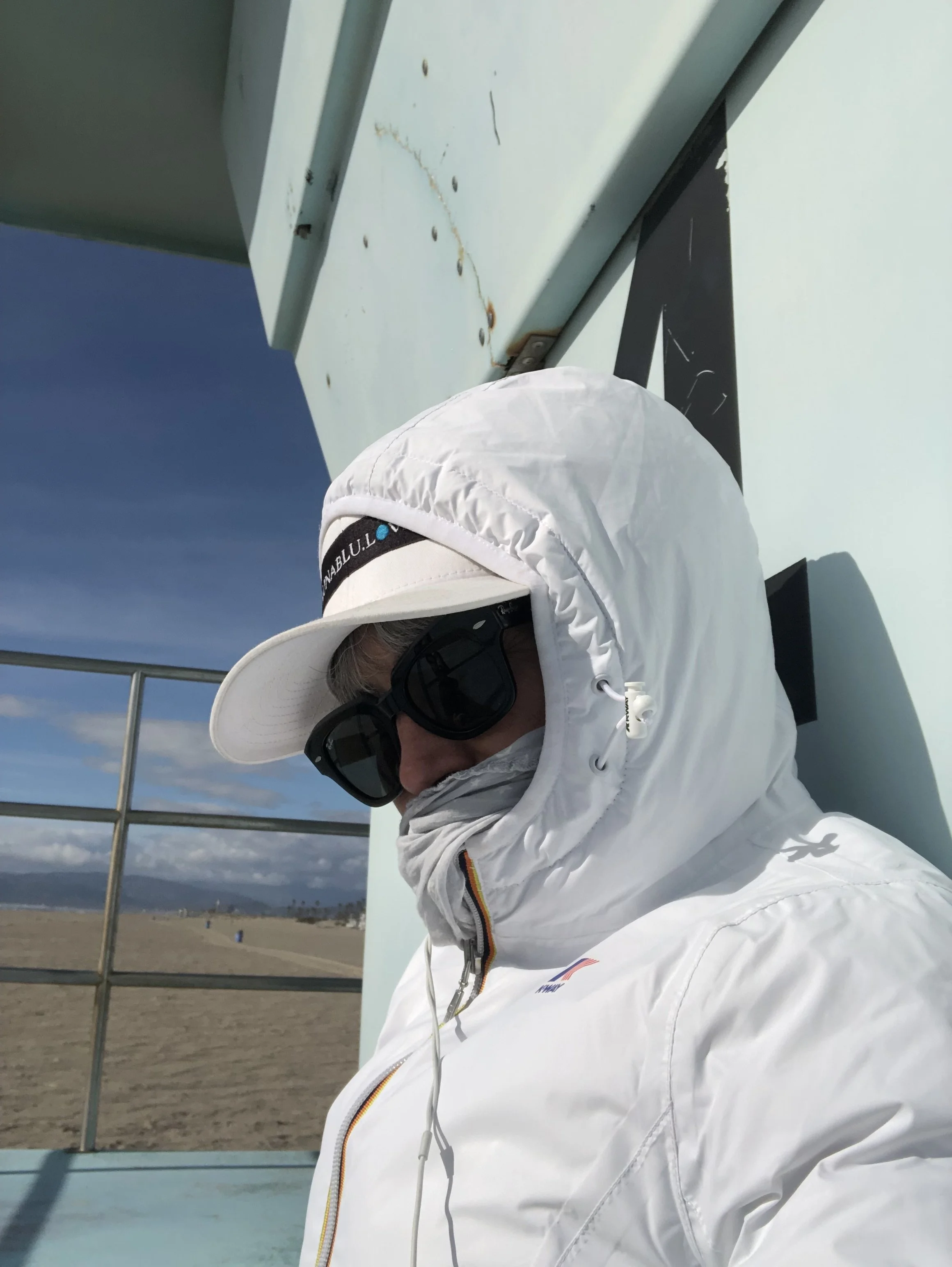 A person wearing a white jacket, large sunglasses, and a white wide-brimmed hat with a black strap. They are standing outdoors near a light green structure with some rust and dirt. The sky is blue with some clouds, and a beach or sandy area is in the