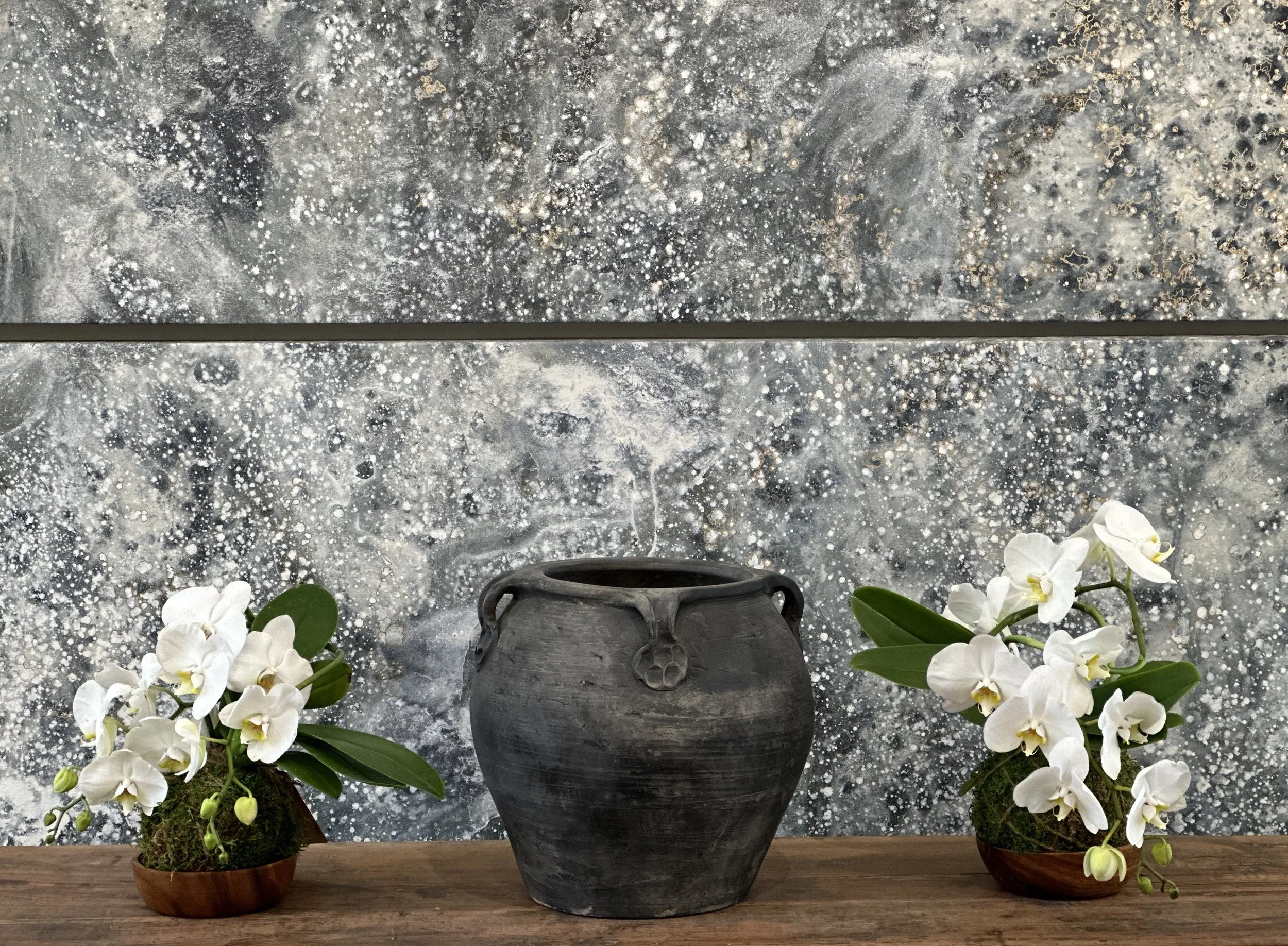 A decorative display with three potted orchids and a dark gray vase on a wooden surface, with a textured gray and white abstract background.