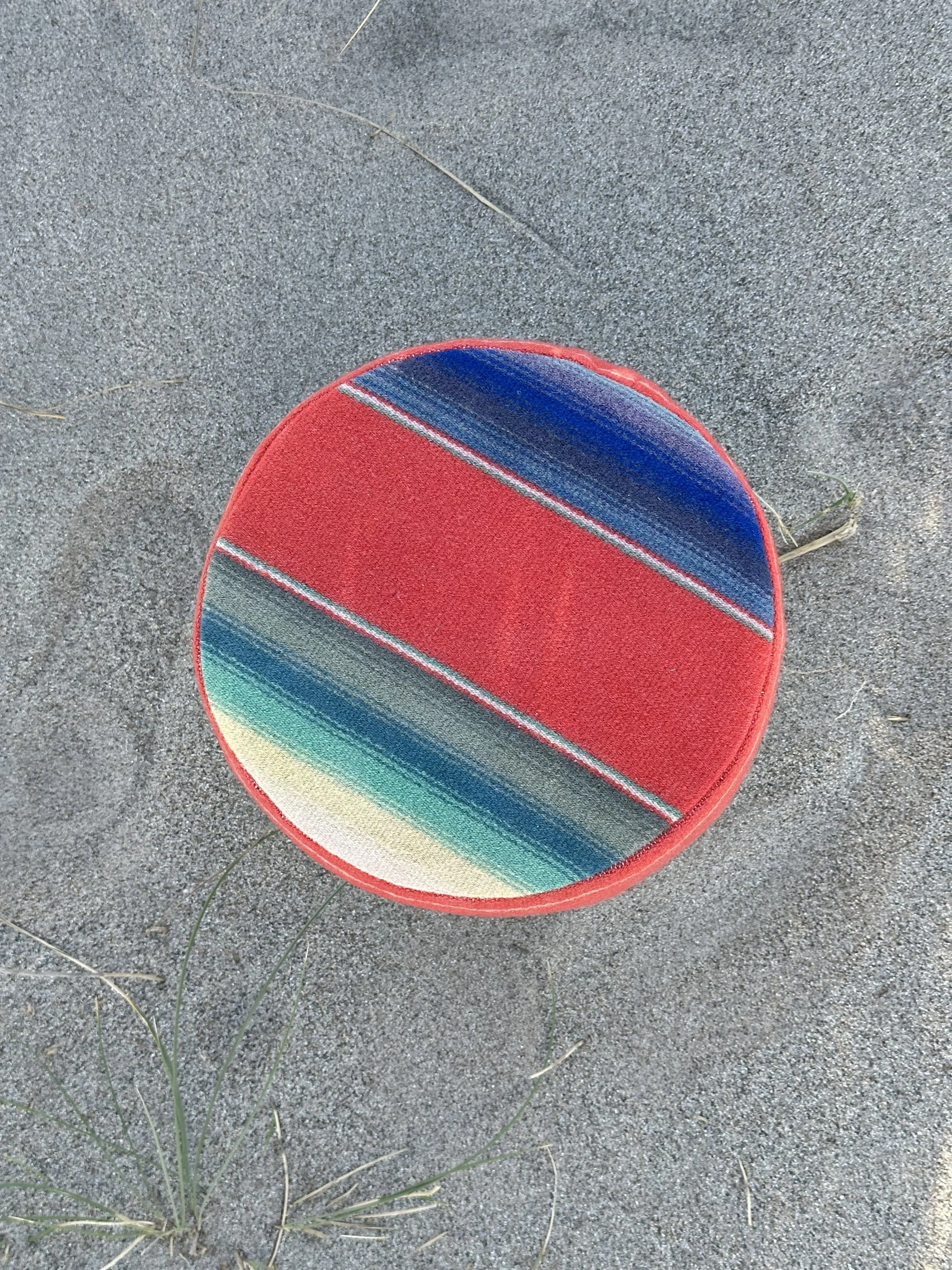A colorful round beach towel with rainbow stripes, lying on sandy ground with some grass and small twigs nearby.