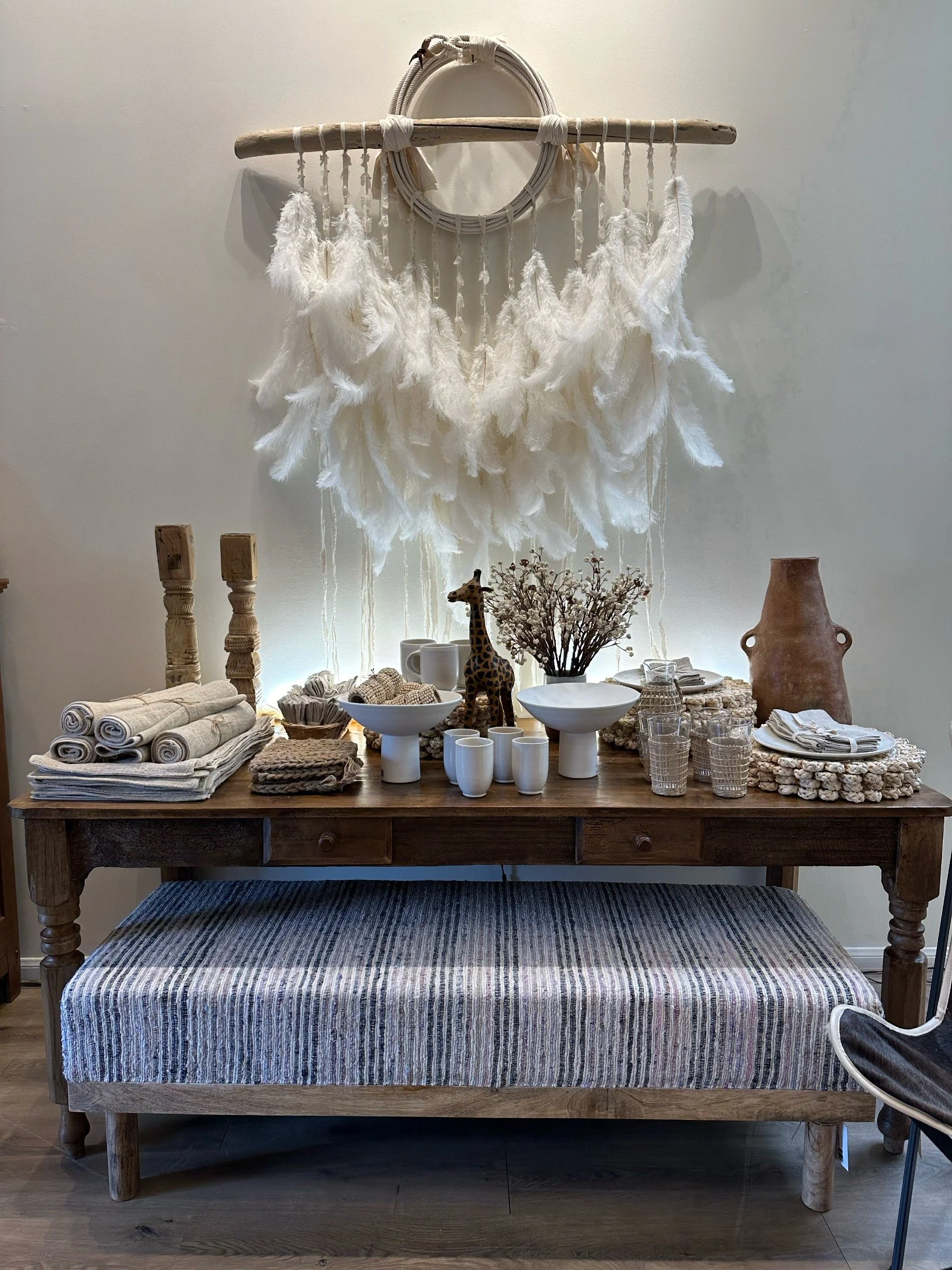 Decorative display on a wooden table featuring white textiles, ceramics, a giraffe figurine, dried flowers, and a large terracotta vase, with a wall hanging of white feathers and circular decor in the background.