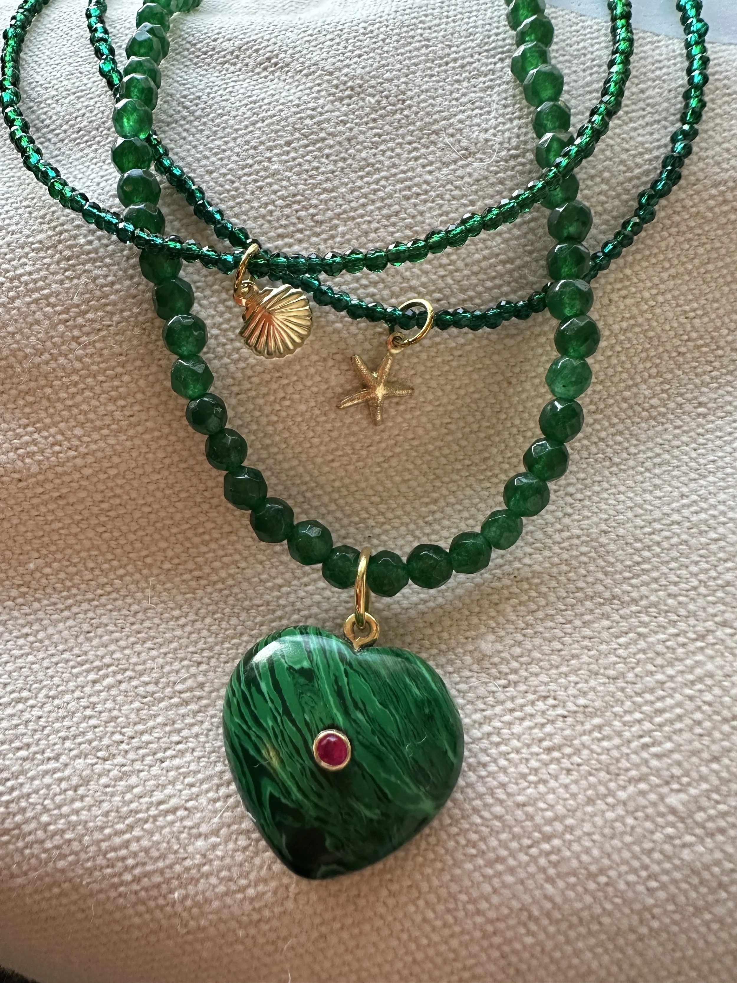 Close-up of a green necklace with a large green pendant, gold and red accents, and smaller green beads on a textured beige background.