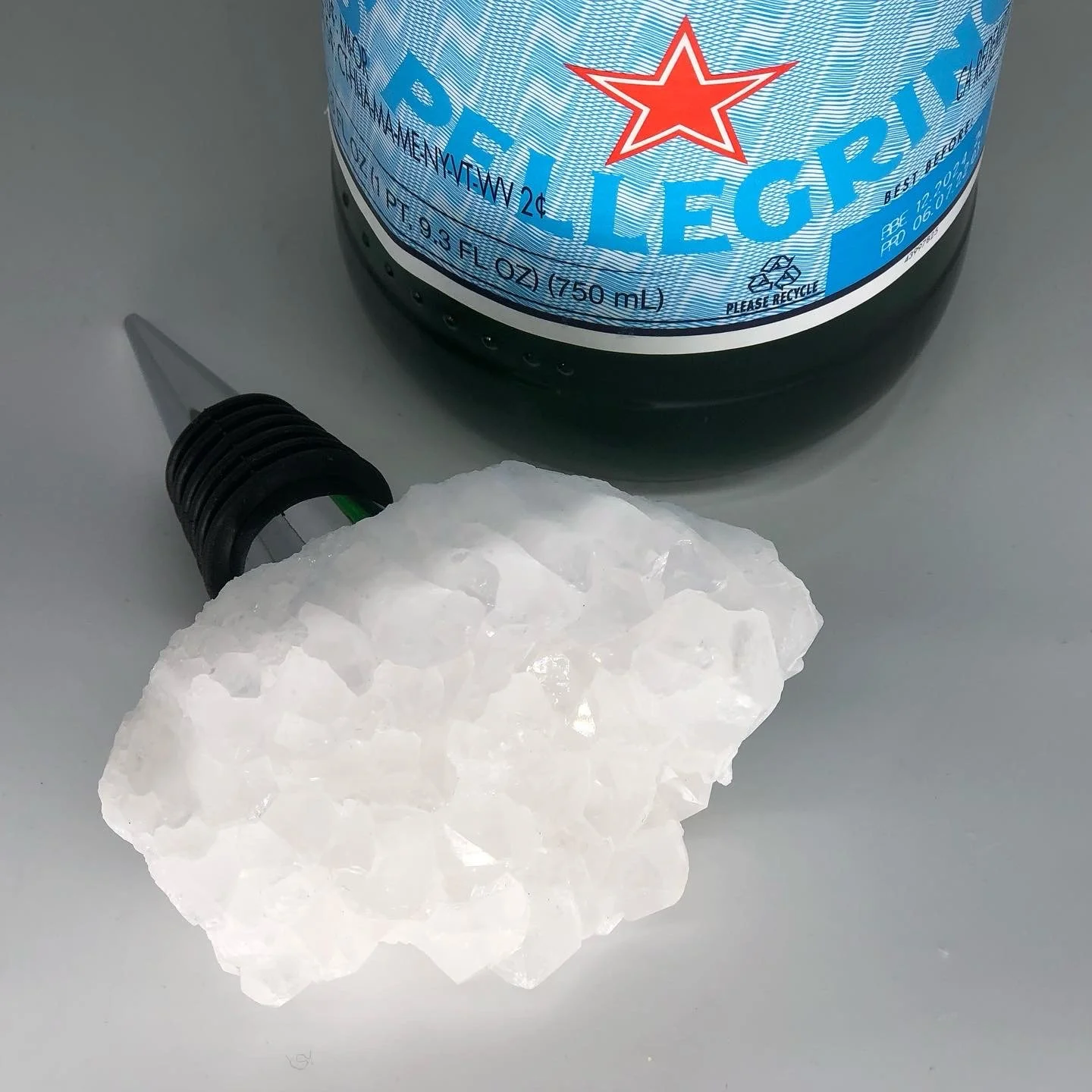 A bottle of Blue Riptide Energy Drink with a black cap and a white and blue label, a piece of white rock salt, and a black flashlight with a green band on a light gray surface.