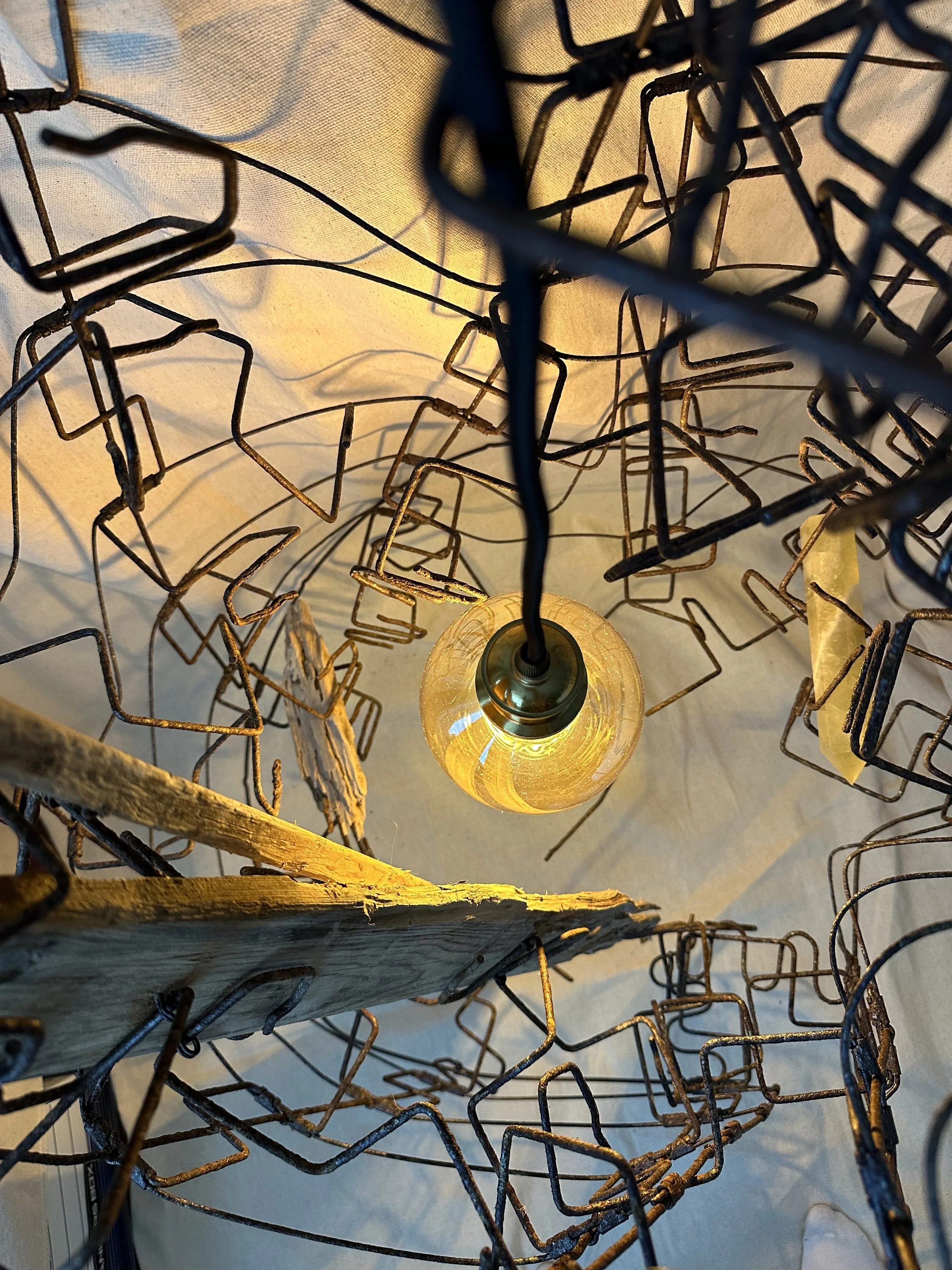 Close-up of a light bulb inside a wireframe lampshade, with a glowing yellowish light. The wireframe is made of black metal and has a rustic, vintage appearance, combined with a piece of wood at the bottom of the frame.