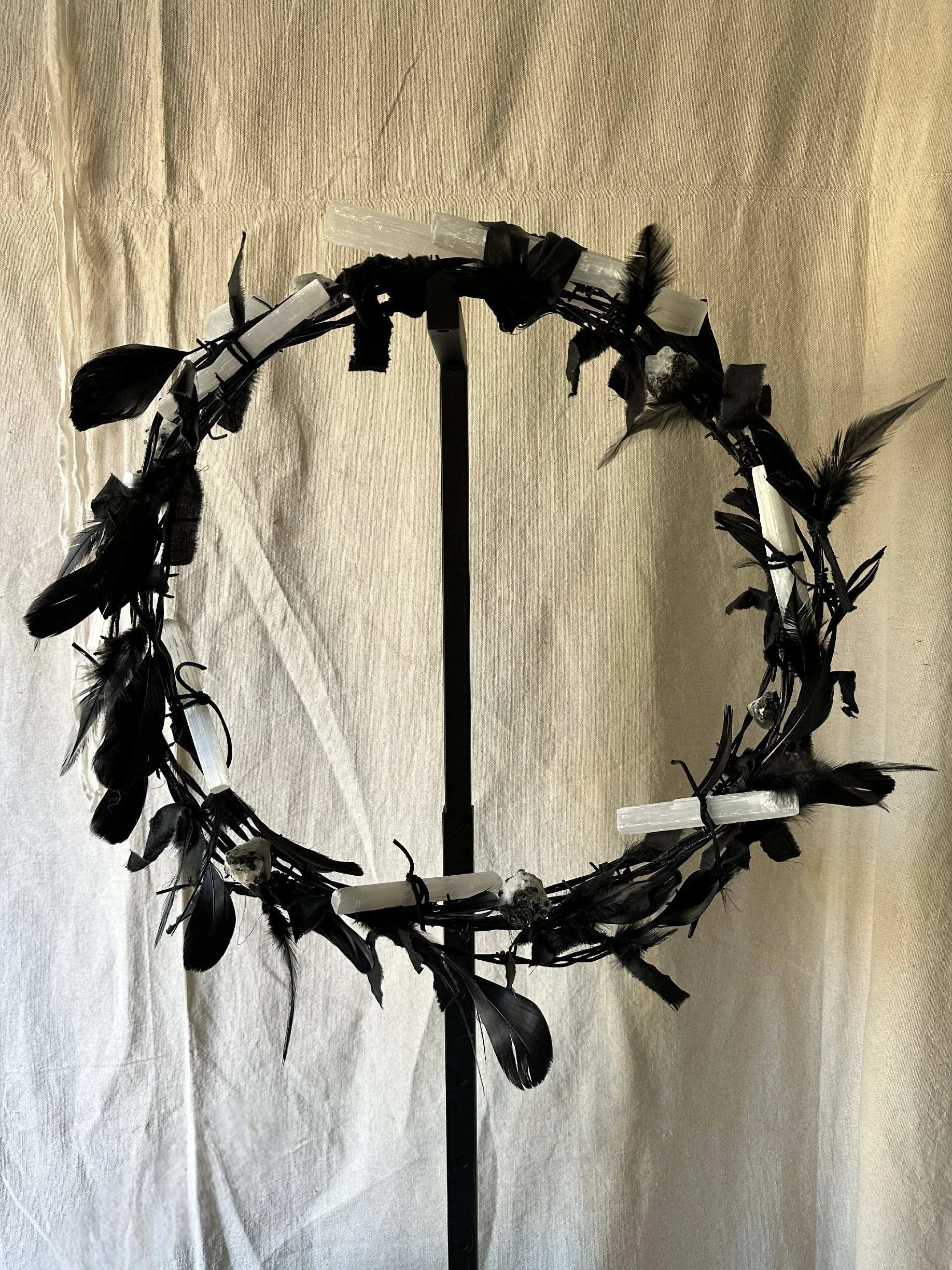 A wreath made of black feathers, white candles, and stones, displayed on a black stand against a beige background.