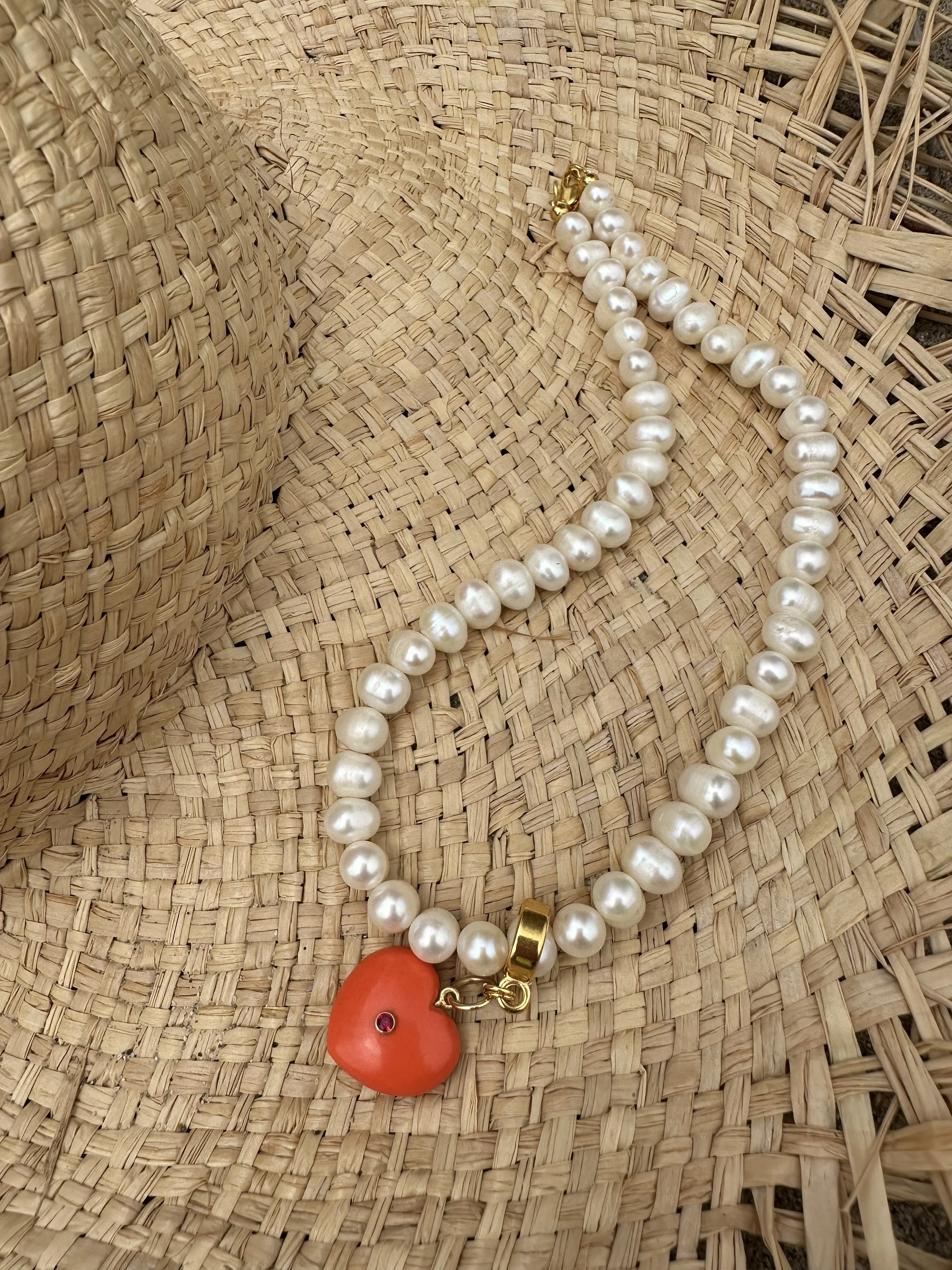 A straw woven handbag with a pearl necklace, a red item with a jewel, and a gold heart-shaped charm placed on it.