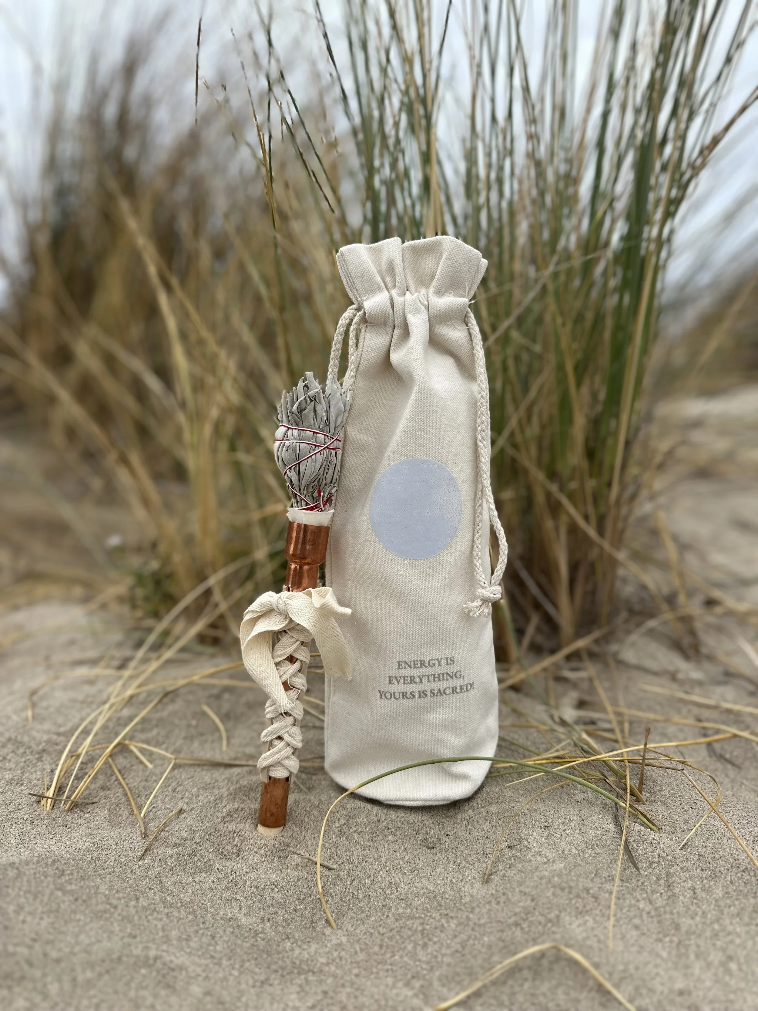 A small drawstring cloth bag hanging on a stick, with a bundle of sage wrapped with red string attached to the bag, in a sandy area with tall grass and plants in the background.