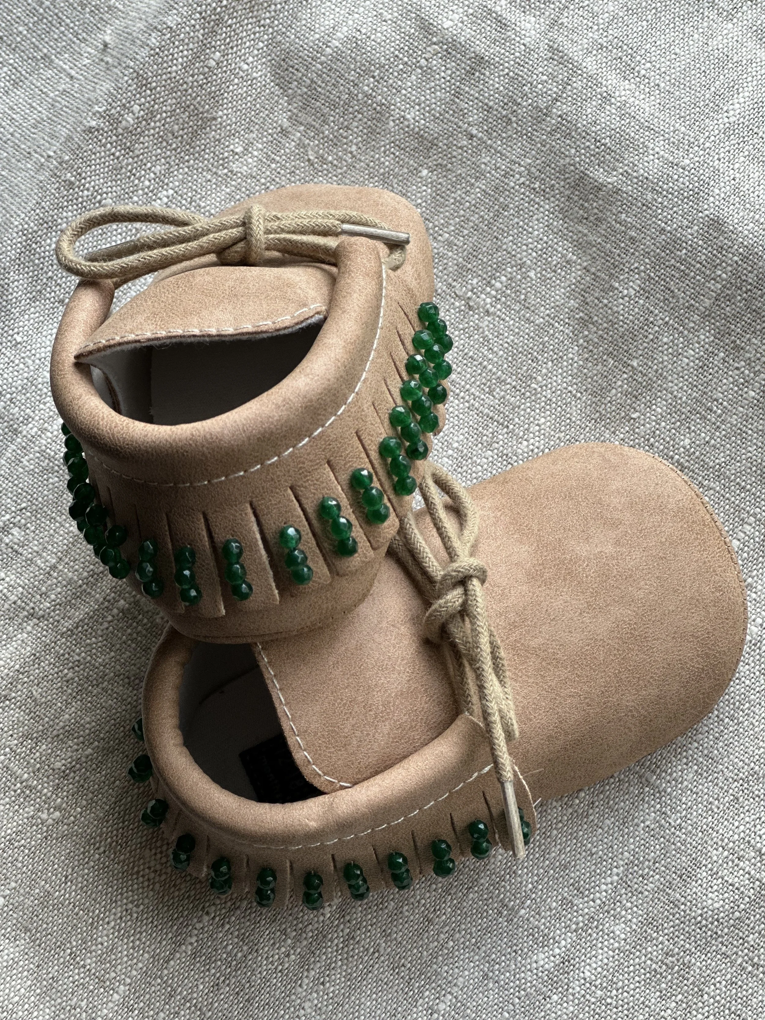 A pair of tan suede sneakers with green bead embellishments on the sides, placed on a textured gray fabric surface.