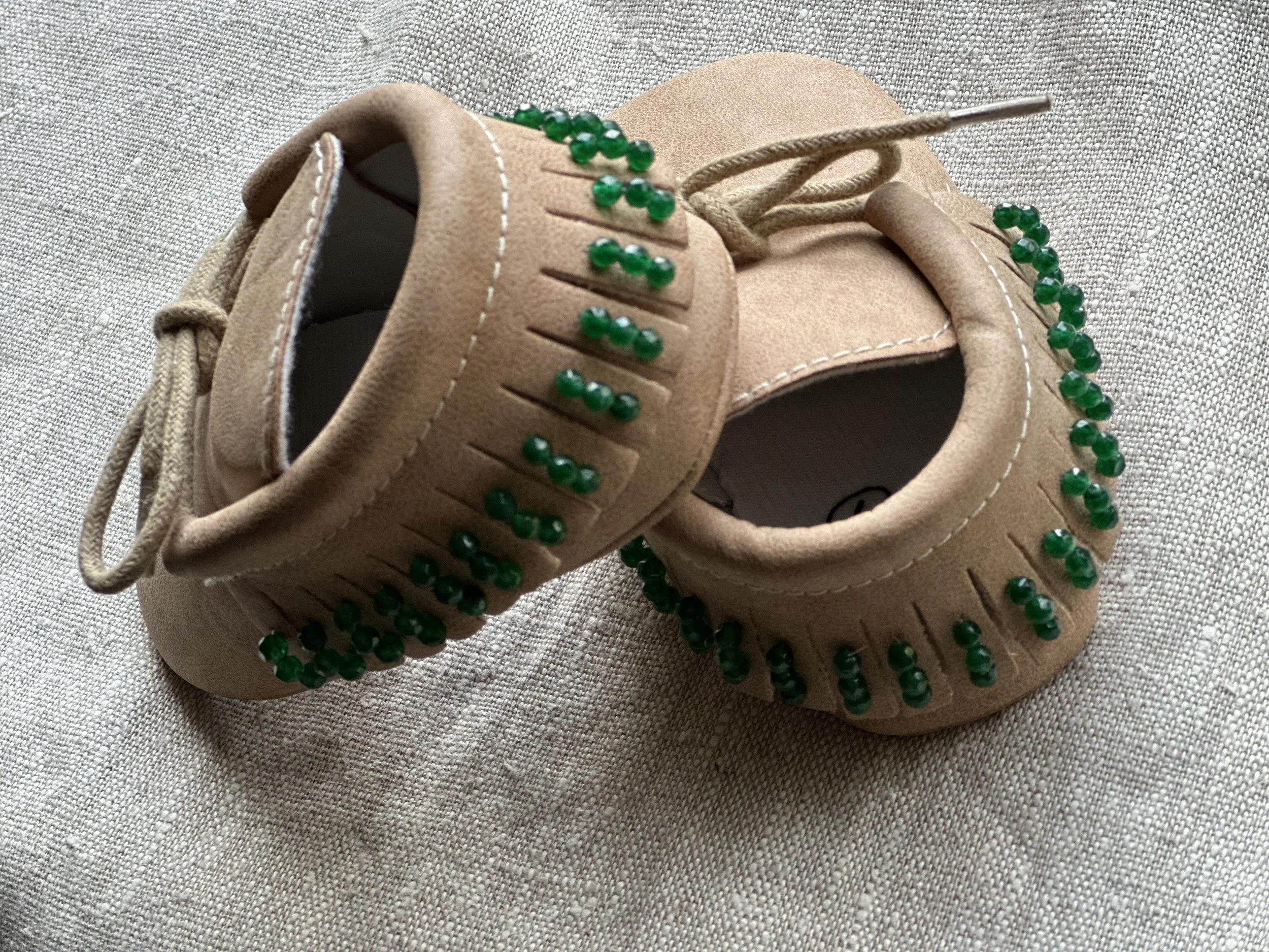 A tan suede sneaker with green bead embellishments on the laces and around the shoe, placed on a light-colored fabric surface.
