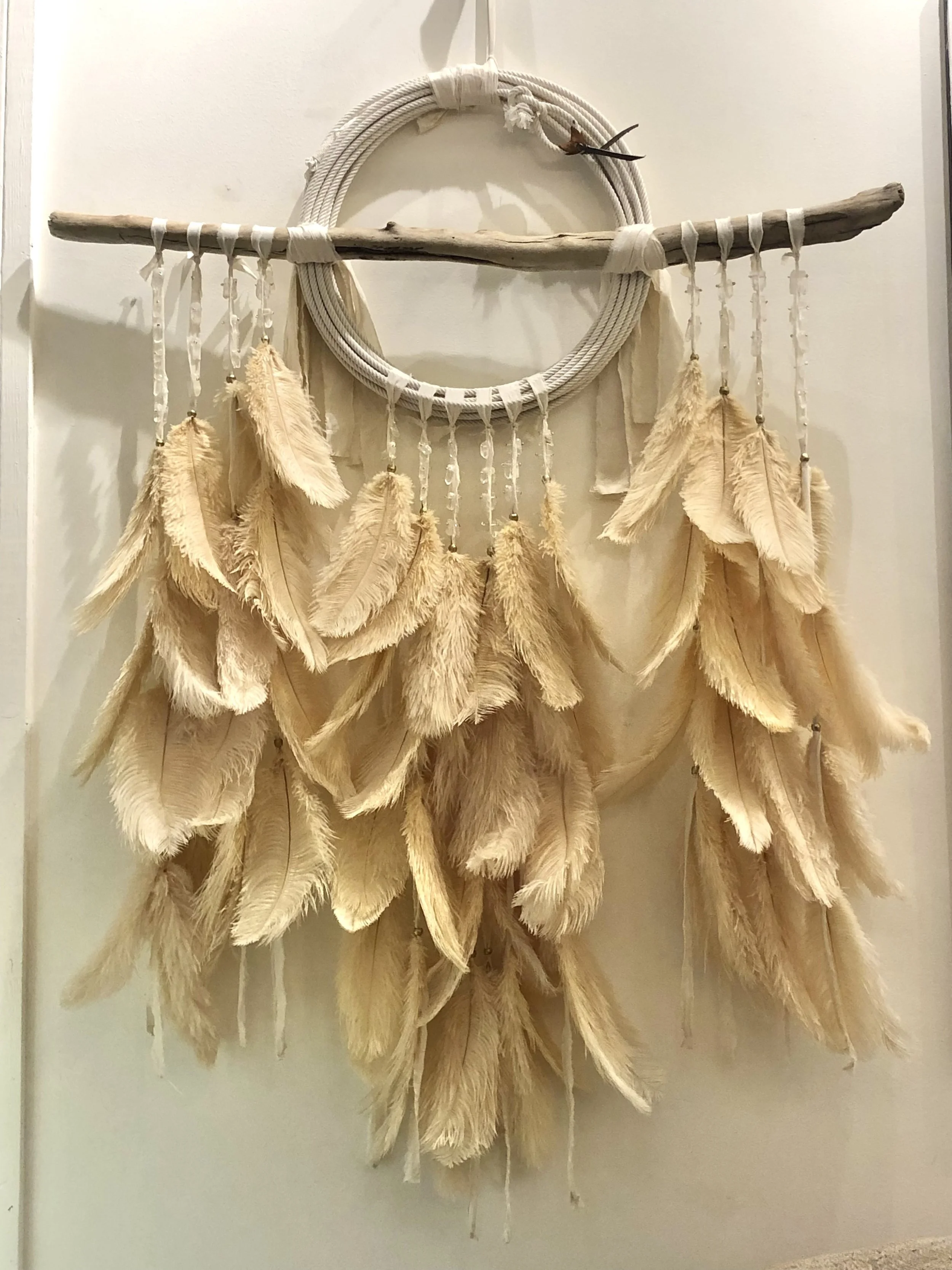 A wall hanging decorative piece featuring a large circular woven ring, a horizontally placed driftwood stick, beige and cream feathers hanging from strings, and a small bird perched on the ring.