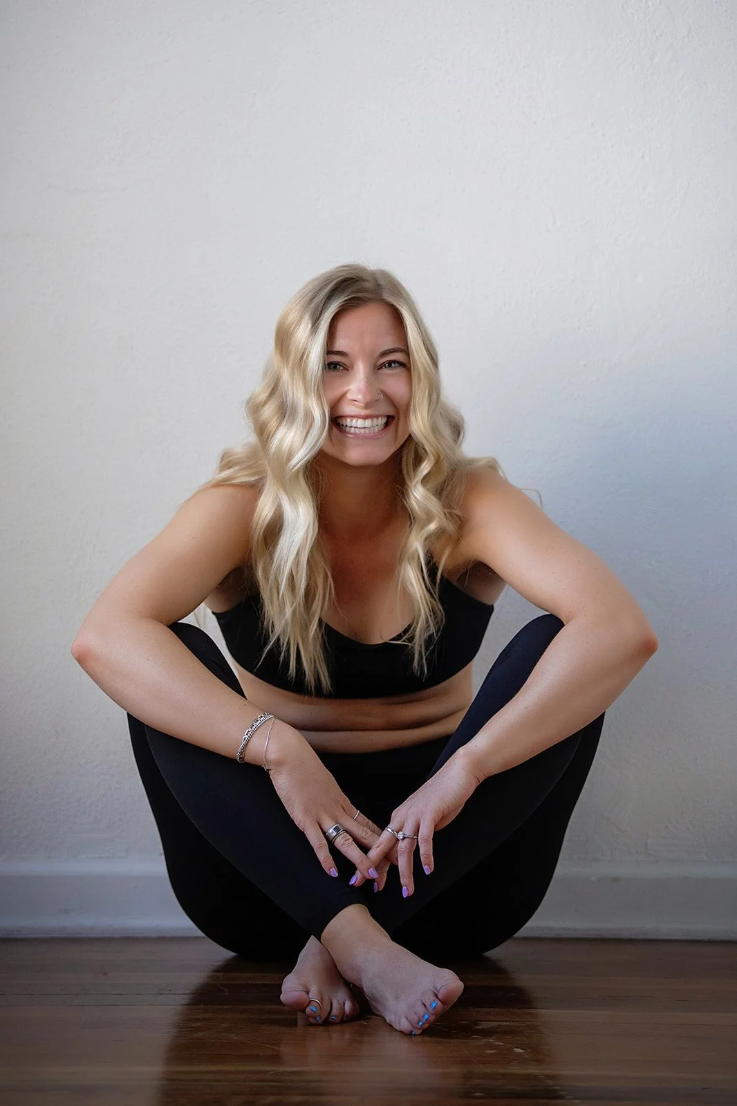 A young woman with long blonde hair, smiling and sitting barefoot on a wooden floor against a plain wall. She is wearing a black sports bra and black leggings, with her arms resting on her knees.