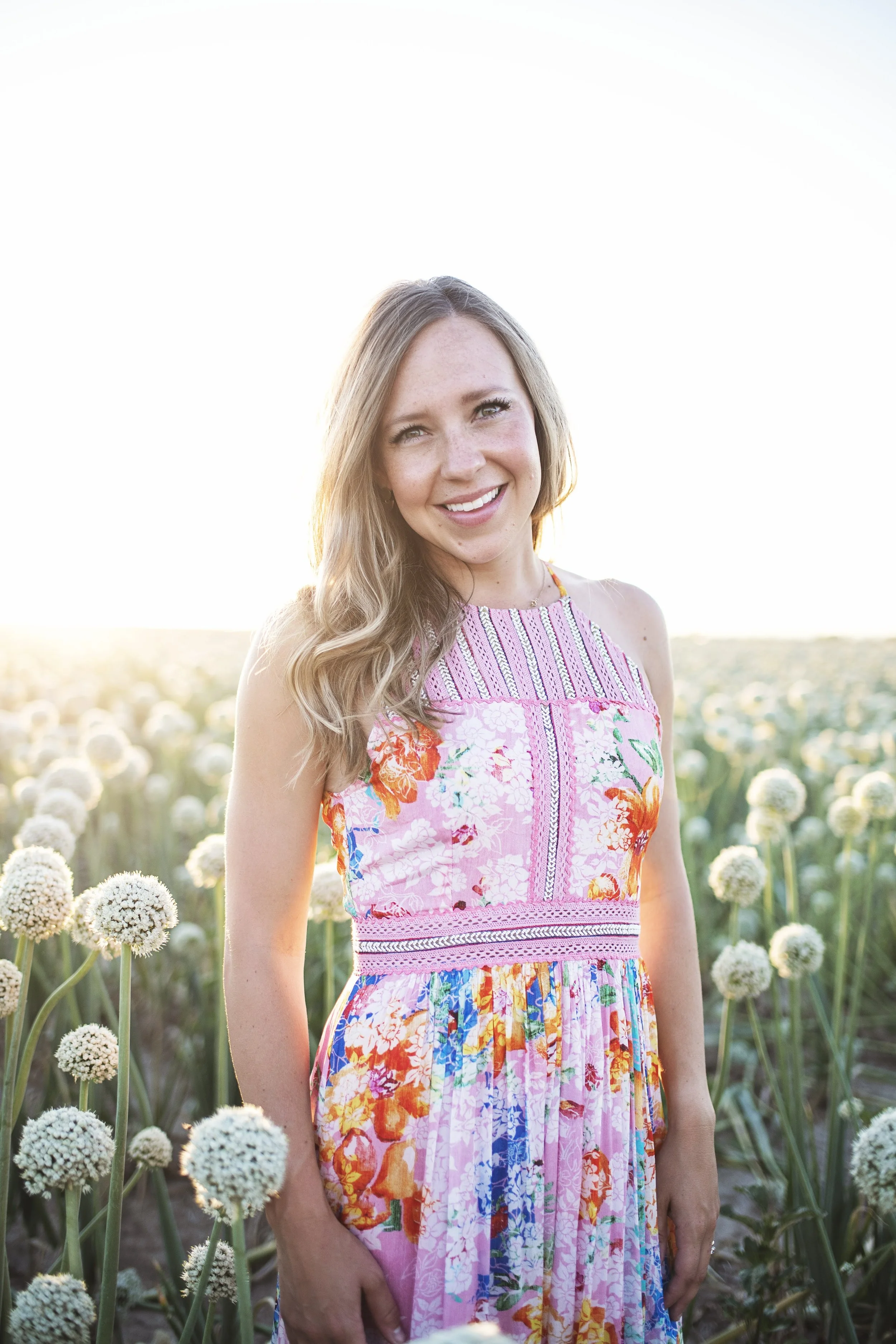 Woman in floral dress standing in a field of flowering plants, smiling with sunlight in the background.