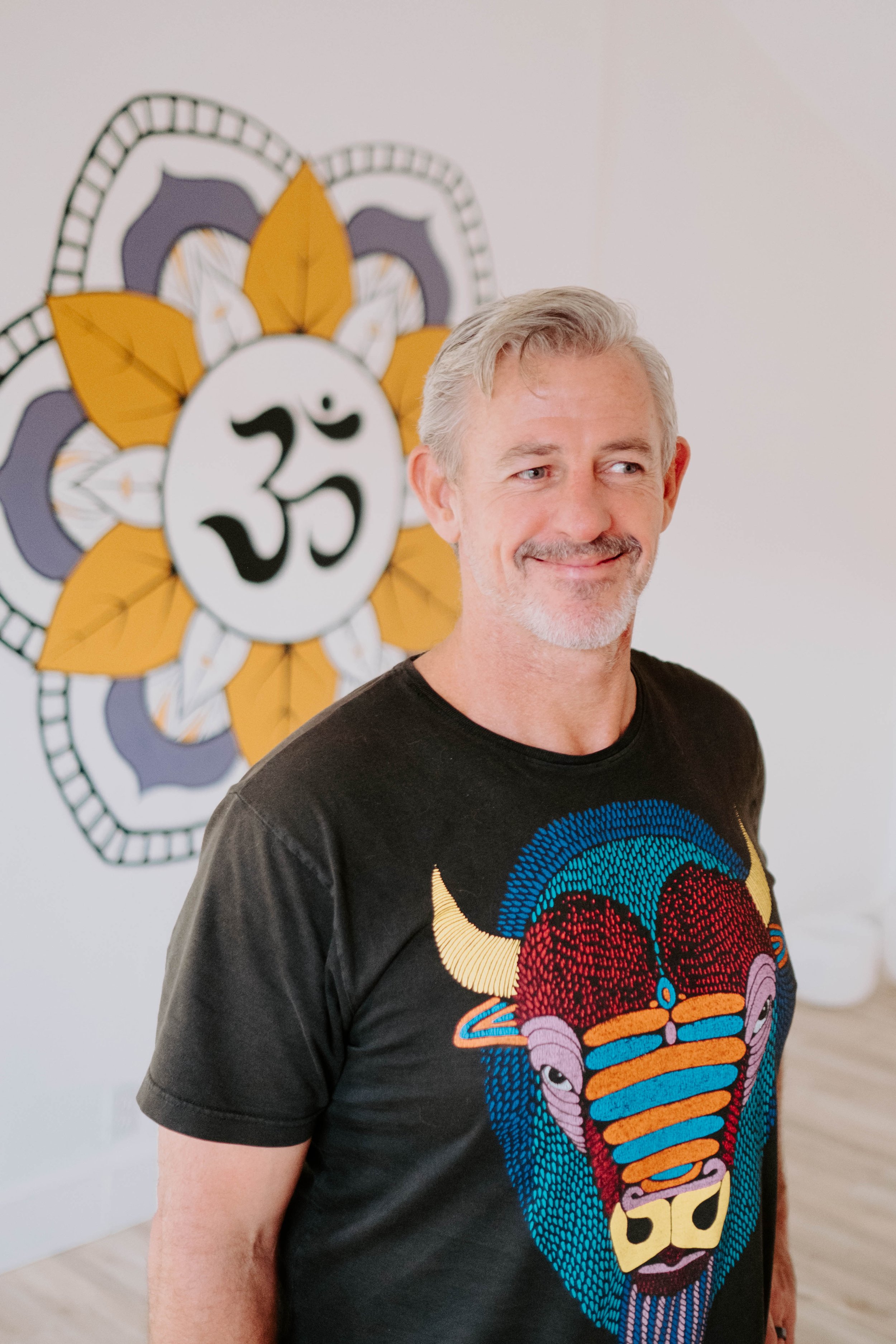 Man with gray hair and mustache wearing a black T-shirt featuring a colorful buffalo design, standing in front of a wall with a yellow and purple mandala containing the Om symbol.