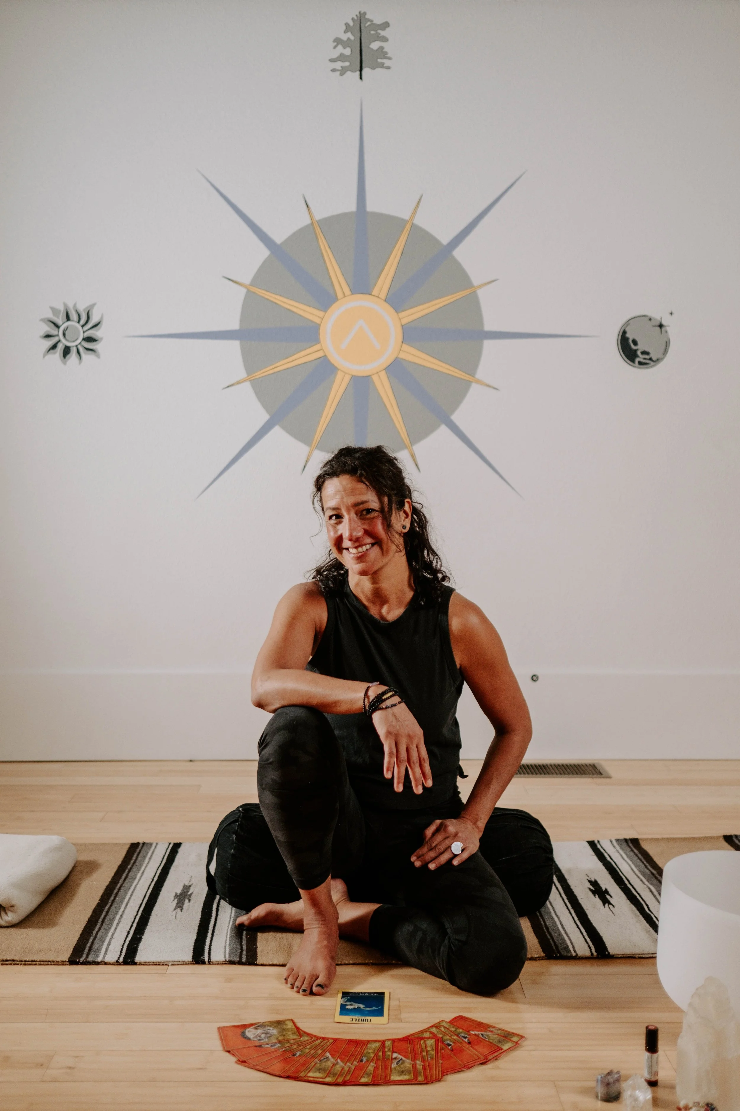 A woman sitting cross-legged on a woven mat in a yoga studio, smiling. Tarot cards, crystals, and a small bottle are placed in front of her. The wall behind has a celestial-themed circular mural.