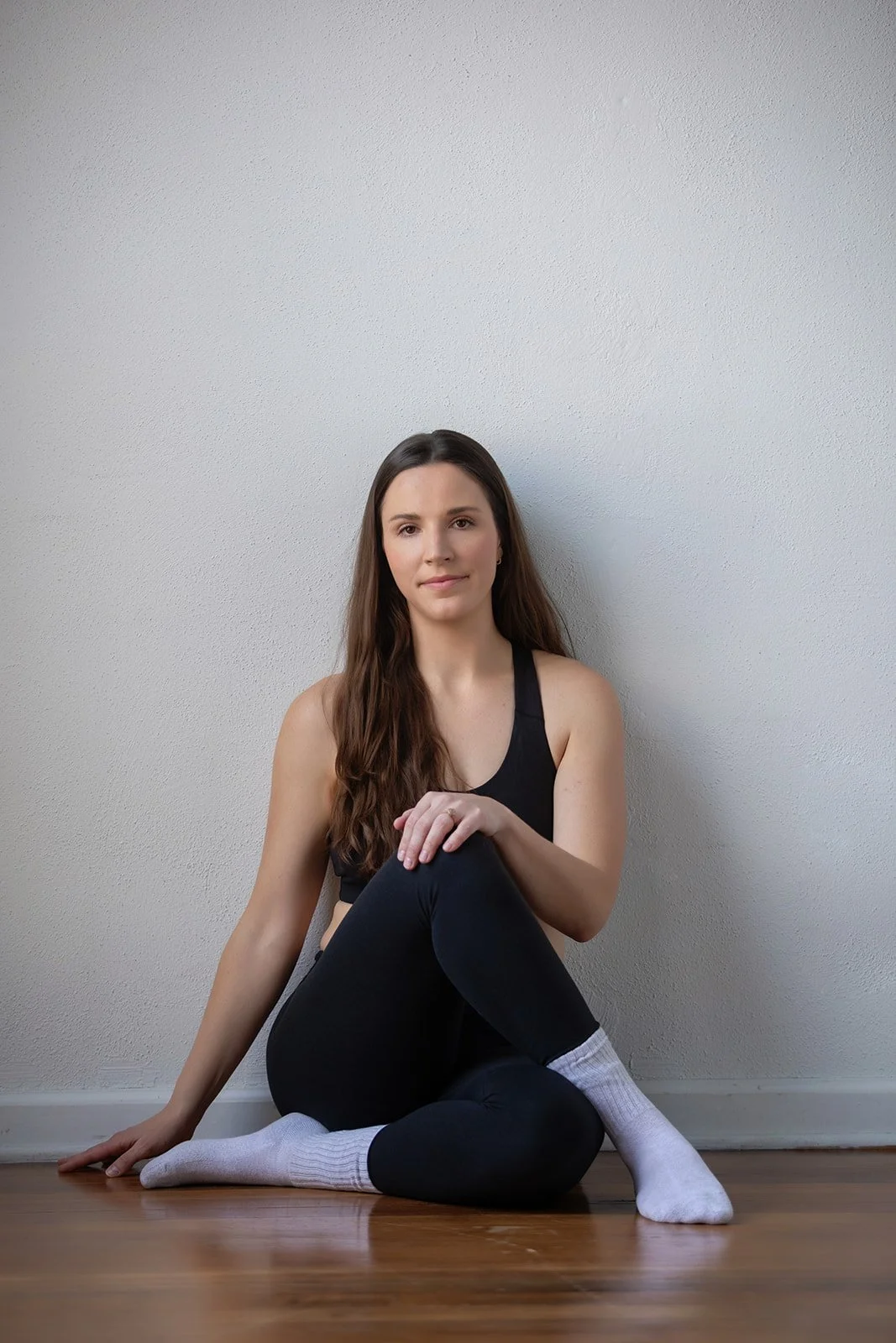 A young woman with long brown hair sitting on a wooden floor against a plain white wall, wearing a black athletic top, black leggings, and white socks, looking at the camera with a neutral expression.