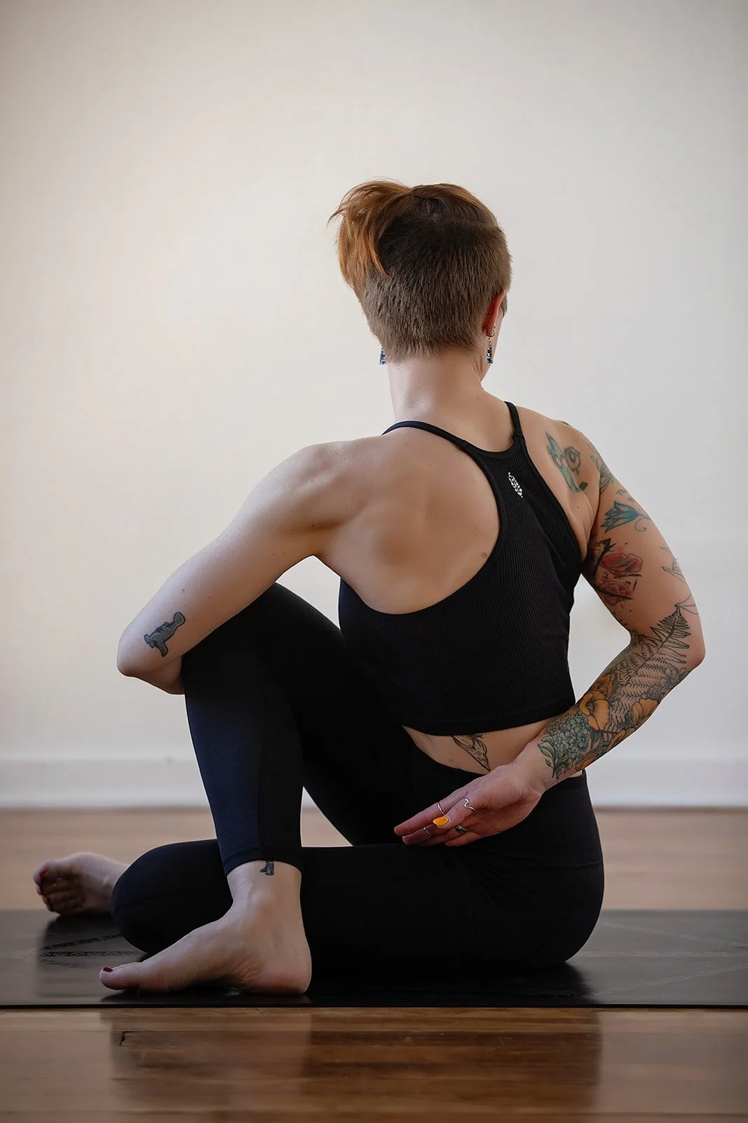 A person practicing yoga in a seated twist pose on a wooden floor, wearing a black sports top and leggings, with short hair and colorful tattoos on both arms.