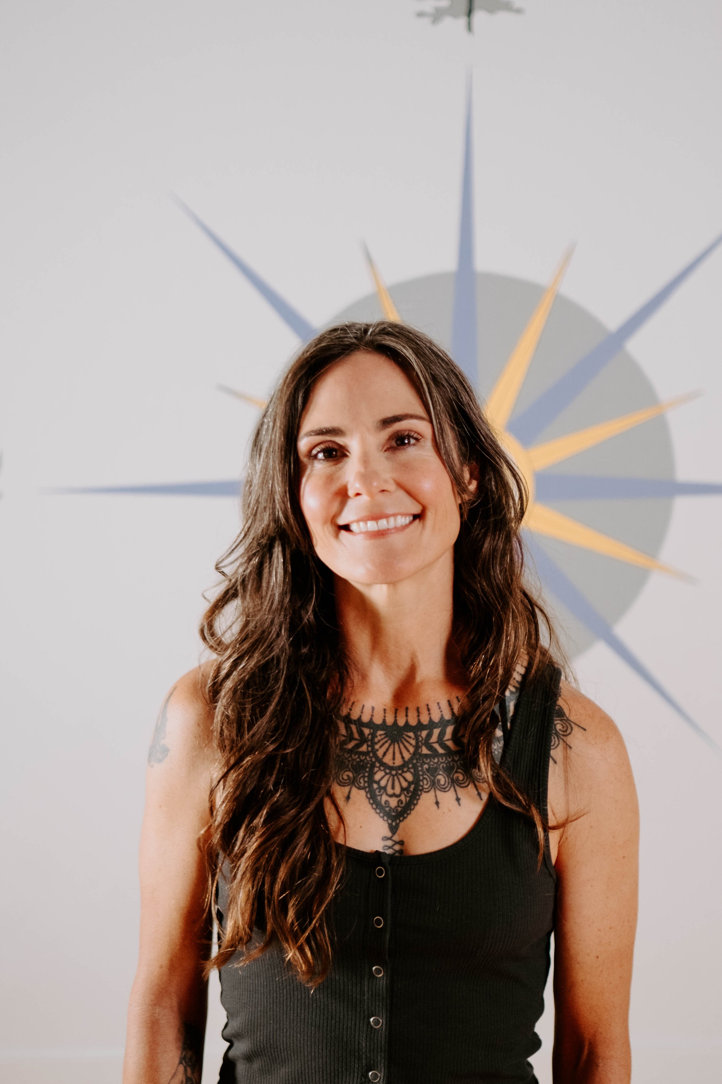 Person with long hair smiling, wearing a dark sleeveless top, and showing tattoos, standing in front of a starburst design on the wall.